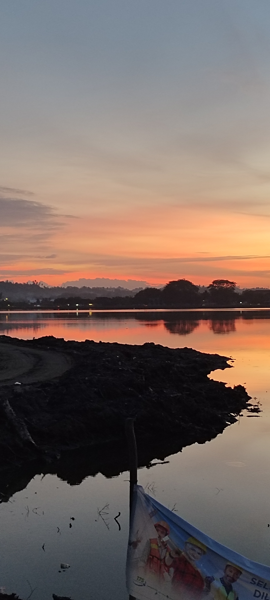 The image captures a serene sunset over a body of water, likely a lake or reservoir, in Lhokseumawe, Indonesia. The sky is a canvas of soft oranges and yellows, transitioning to a muted blue at the top, indicating the approach of twilight. Silhouetted against the vibrant sky are rolling hills and scattered trees, forming a distant horizon line. The water's surface acts as a mirror, reflecting the colorful sky and the dark silhouettes of the land, creating a symmetrical and peaceful scene.
In the foreground, a pile of dark, coarse material, possibly soil or construction debris, juts into the water, creating a stark contrast with the calm water and the colorful sky. The texture of this material is rough and uneven, suggesting a natural or recently disturbed environment. Partially submerged in the water and leaning against a wooden pole is a large, blue and white banner. The banner features images of three men in construction attire, including hard hats and safety vests, with text that appears to be in Indonesian. The presence of the banner and the construction debris suggests that this area might be undergoing some form of development or infrastructure work within or near Lhokseumawe. The overall impression is one of natural beauty juxtaposed with ongoing human activity.
