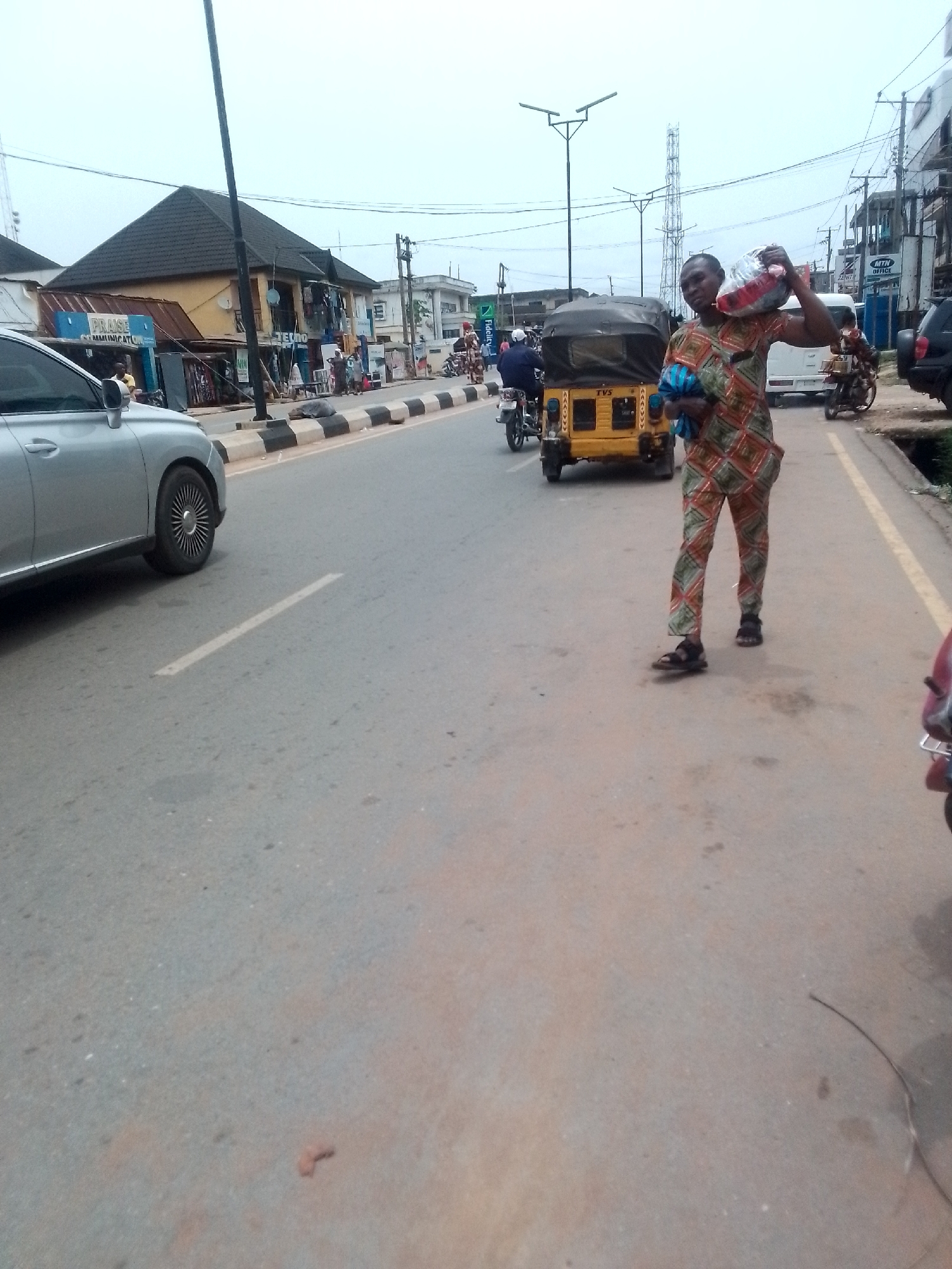 A street in Orlu, Nigeria, features a man carrying a large bag on his shoulder. He is wearing a patterned orange and green outfit with sandals. Beside him on the road is a yellow tricycle. Other vehicles, including cars and motorcycles, are on the street, and pedestrians are visible in the background near shops and buildings. Streetlights and utility poles line the road.