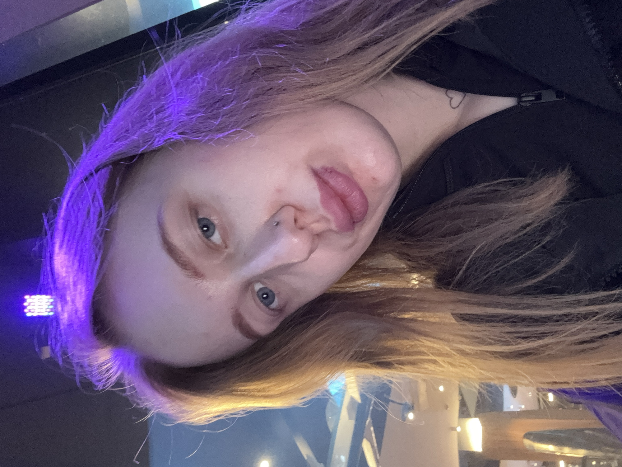 A fair-skinned individual with dyed blonde hair is facing the camera with their tongue extended. They have a pierced nose and blue eyes. A black zipper jacket is visible. A small heart tattoo can be seen on their neck. The background appears to be a brightly lit, possibly public space with blurred elements. The image is taken at an angle. There is no indication this image was taken in Ol'ginskaya, Russia.