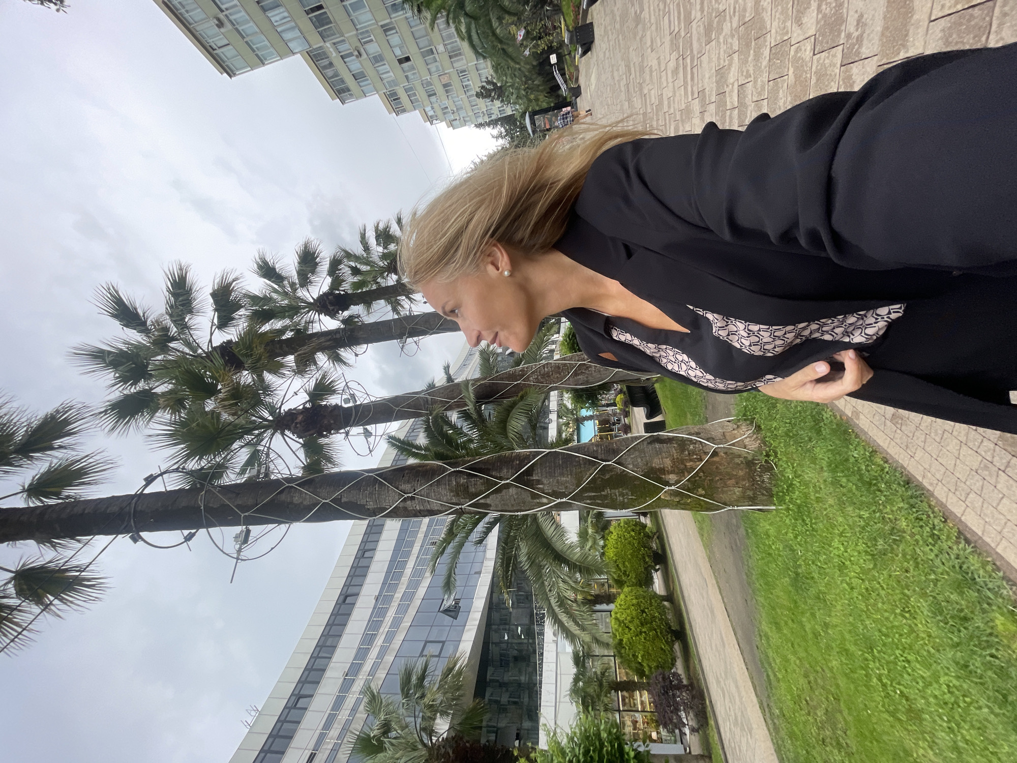 A woman with blonde hair, wearing a black blazer and a black top with a patterned neckline, stands on a brick-paved walkway next to a grassy area. Palm trees line the walkway, and some are encased in a white netting. A modern building with reflective windows rises in the background, along with other tall buildings and a cloudy sky. This scene is located in Sochi, Russia.