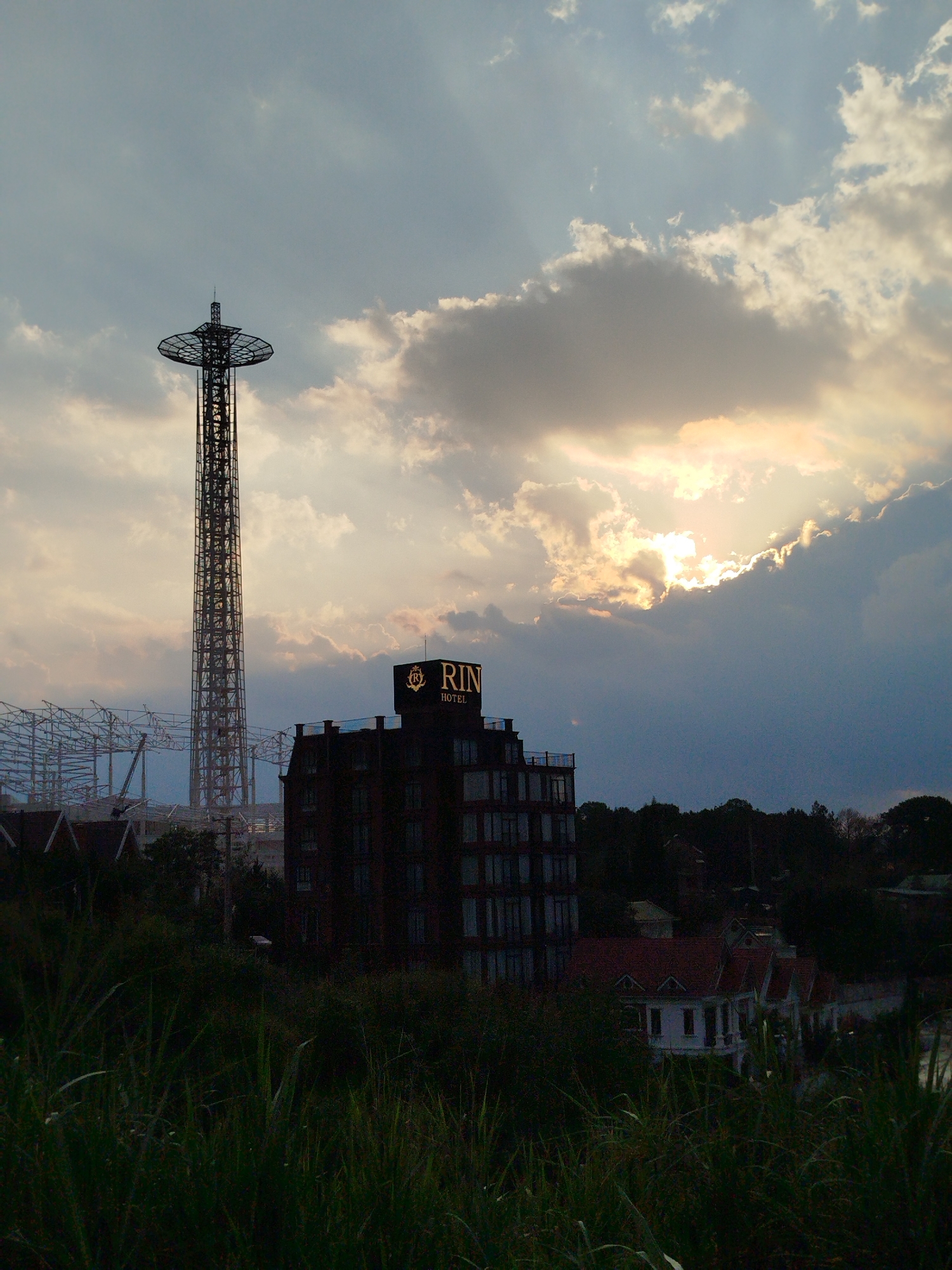 This image shows a cityscape in Da Lat, Vietnam, featuring a tall, open-lattice tower and the Rin Hotel. The tower is silhouetted against a cloudy sky with the sun peaking through, creating crepuscular rays. The Rin Hotel, a multi-story building, is also silhouetted, with its illuminated sign visible. In the foreground, overgrown grass and foliage obscure the view of a white house with a red-tiled roof. In the background, there is construction with a metal lattice framework. The overall scene appears to be taken during sunset or sunrise.
