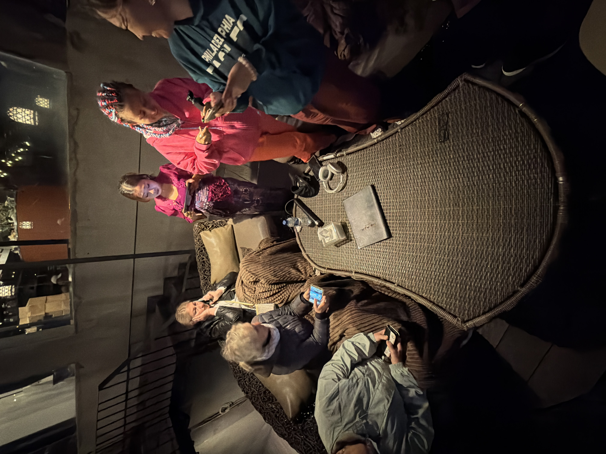 The image depicts a group of six individuals gathered on a patio or rooftop seating area at night. The setting appears to be outdoors, possibly a restaurant or bar, with ambient lighting from overhead fixtures and possibly distant city lights visible in the background through a window. The individuals are mostly focused on their mobile devices.
From left to right, a person in a light blue jacket is seated, holding a phone in their right hand. Next to them, a woman with blonde hair, wearing a dark grey jacket and a scarf, is also seated and looking at her phone. Further back, a woman in a black leather jacket is seated on a couch and appears to be speaking on her phone, with one hand touching her face. In the center, a young girl in a pink dress is standing and looking at her phone. To her right, a woman with colorful braided hair, wearing a pink hooded jacket and orange pants, is also looking at her phone. On the far right, a person wearing a teal sweatshirt with "PHILADELPHIA" printed on it is also engaged with their phone.
In the foreground, a woven rattan coffee table holds several items, including a laptop, a small notebook, a bottle of water, and a pair of headphones. The overall atmosphere is casual and modern. The background shows architectural elements like a staircase and a window with visible shelving and decorative lights. The location in Kintamani, Indonesia, is not directly evident from the visual cues in the image.