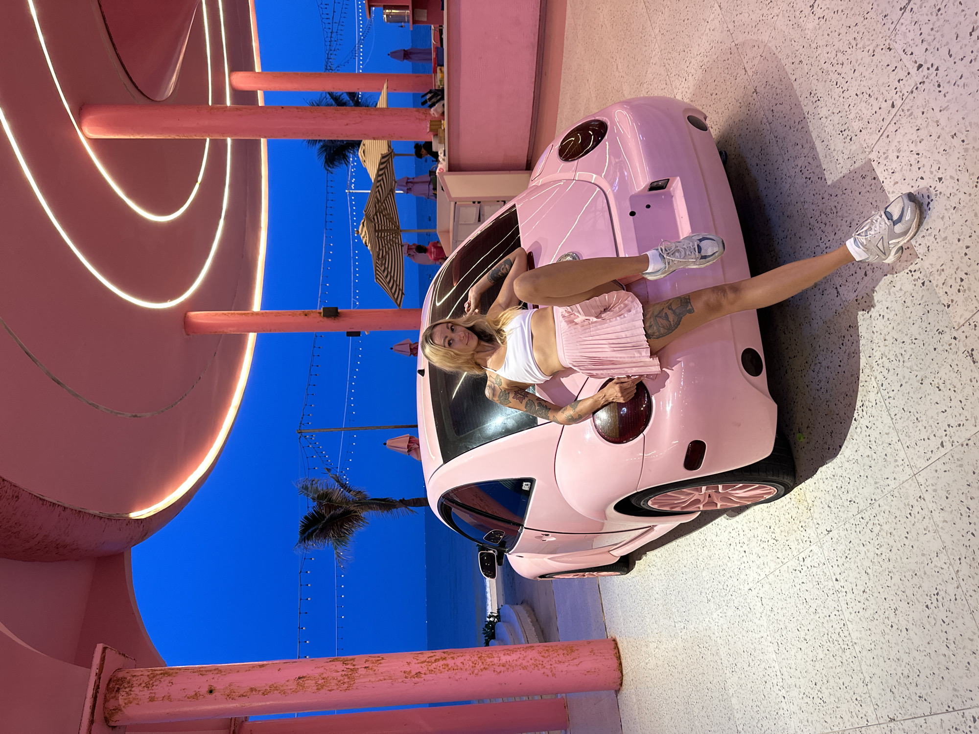 A young Caucasian woman with blonde hair and tattoos is sitting on the hood of a pink Volkswagen Beetle. She is wearing a white sports bra and a pink pleated skirt, with white sneakers. The car is parked on a speckled gray tiled patio. In the background, the ocean can be seen under a deep blue twilight sky. The scene is framed by tall, pink cylindrical pillars supporting a curved pink roof with embedded lights. In the distance, there are beach umbrellas and a bar area. The location is identified as Tien Thanh Ward, Vietnam.