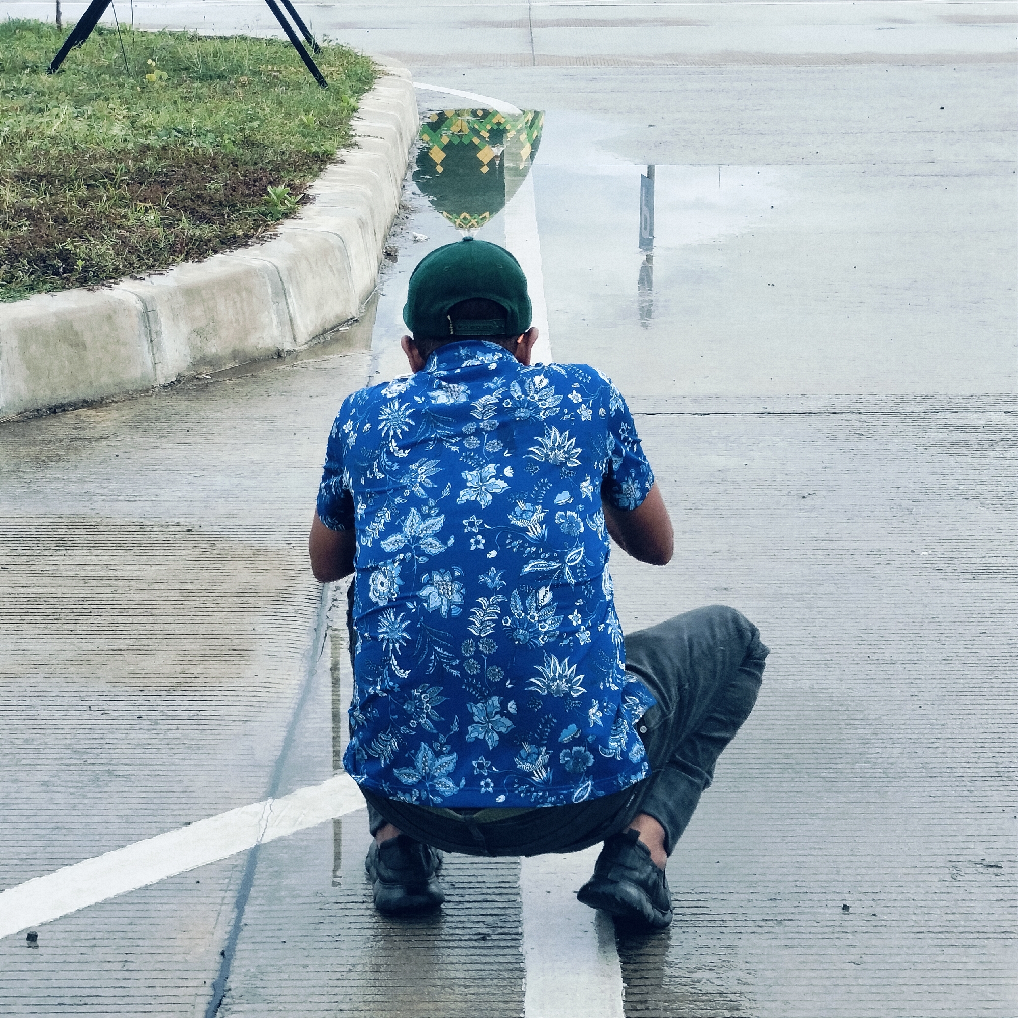 An outdoor, slightly elevated view captures a person squatting on a wet, paved surface, likely a road or large pathway, next to a concrete curb and a strip of grass. The person is seen from the back, wearing a dark green baseball cap, a vibrant blue short-sleeved shirt adorned with a white and light blue floral pattern, and dark trousers or jeans. Their feet are clad in dark athletic shoes, and they appear to be focused downwards, possibly observing something on the wet ground or within the puddles.
The ground is uniformly wet with several puddles, suggesting recent rain or standing water. A clear white painted line runs vertically through the frame, directly beneath the person's squatting position. In a prominent puddle to the right of the person, a distinct reflection shows a green and yellow geometric pattern, reminiscent of architectural designs often found on domes or structures in Islamic cultures, consistent with the Keureuweungkrueng, Indonesia location. Further down the wet pavement, a more distant puddle shows a faint reflection that includes what appears to be the number "6" or a similar numerical character from a sign. The sky appears overcast, contributing to the diffused lighting.
