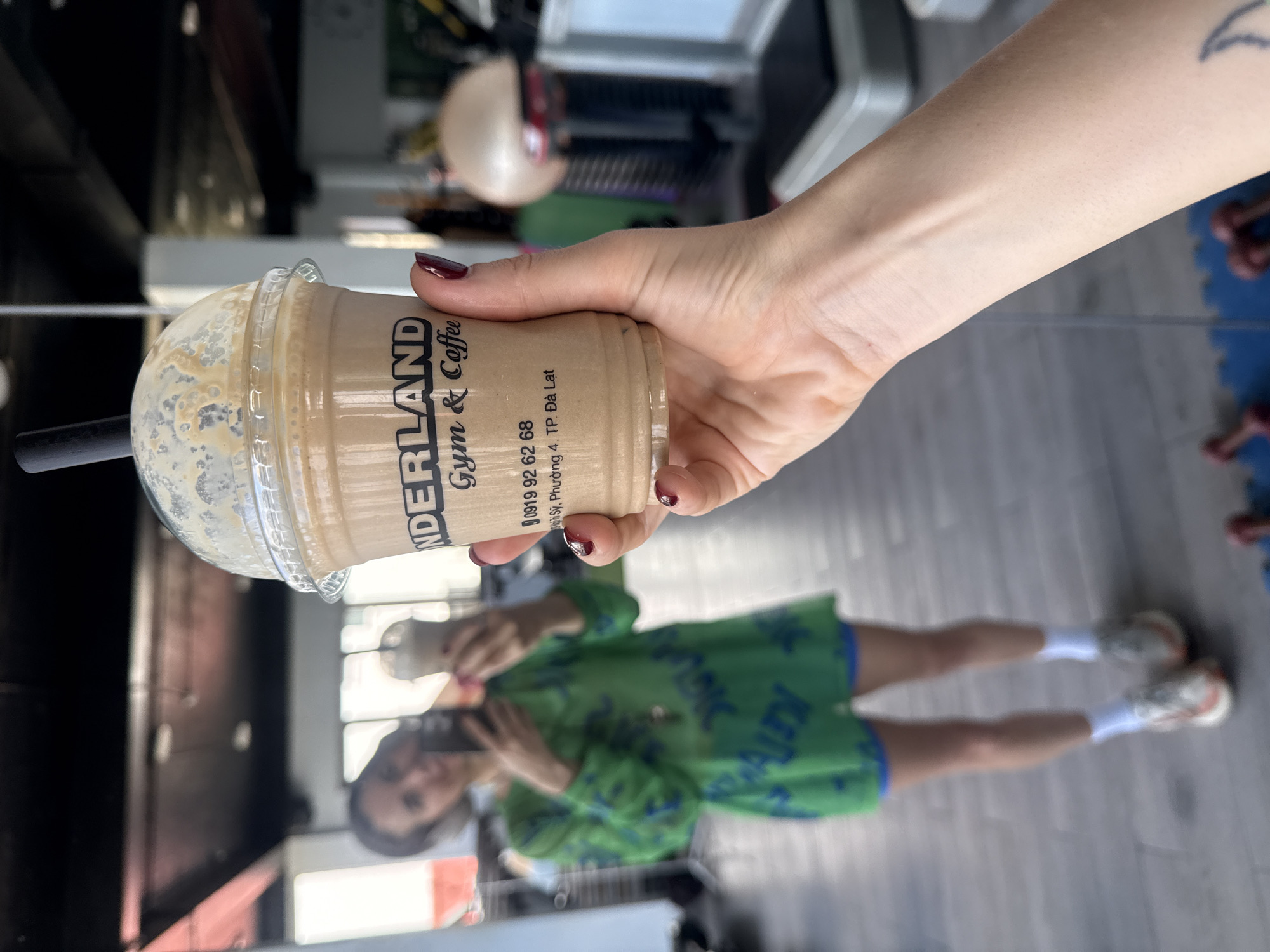 In the foreground, a person's hand with dark red nail polish holds a plastic cup of iced coffee. The cup has a label with the text "NDERLAND Gym & Coffee" and a phone number "0919 92 62 68" and an address "19.Duong 4, TP. Da Lat". In the background, a woman wearing a bright green top and blue shorts is taking a selfie with her phone. She is standing in a gym with exercise equipment visible. The flooring appears to be grey tile.