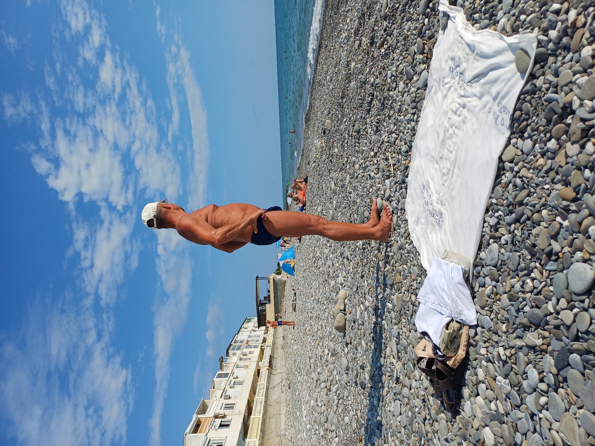 A man in blue swim trunks and a white cap is in the foreground of this image, positioned horizontally. His body is taut as if he is performing a balancing act, with his legs extended and arms held out to his sides. He appears to be on a pebble beach, with the sea stretching out behind him. The water is a calm blue, with small waves lapping at the shore.
In the background, there are several other people on the beach, some of whom are swimming in the sea. A white building with multiple balconies lines the edge of the beach, suggesting a resort or residential area. Several beach umbrellas and a small structure that looks like a cafe are also visible.
The image is taken on a bright, sunny day, as indicated by the clear blue sky and scattered white clouds. The overall mood is relaxed and summery, consistent with a vacation at the beach. The pebbles on the beach are varied in size and color, with some grey and white stones being prominent.
There is some visible text on a white cloth laid out on the pebbles, but it is not clear enough to be legible. The context provided indicates the location is Sochi, Russia, which is a coastal city known for its beaches and resorts. The time of day appears to be daytime, likely in the morning or afternoon, given the bright sunlight.