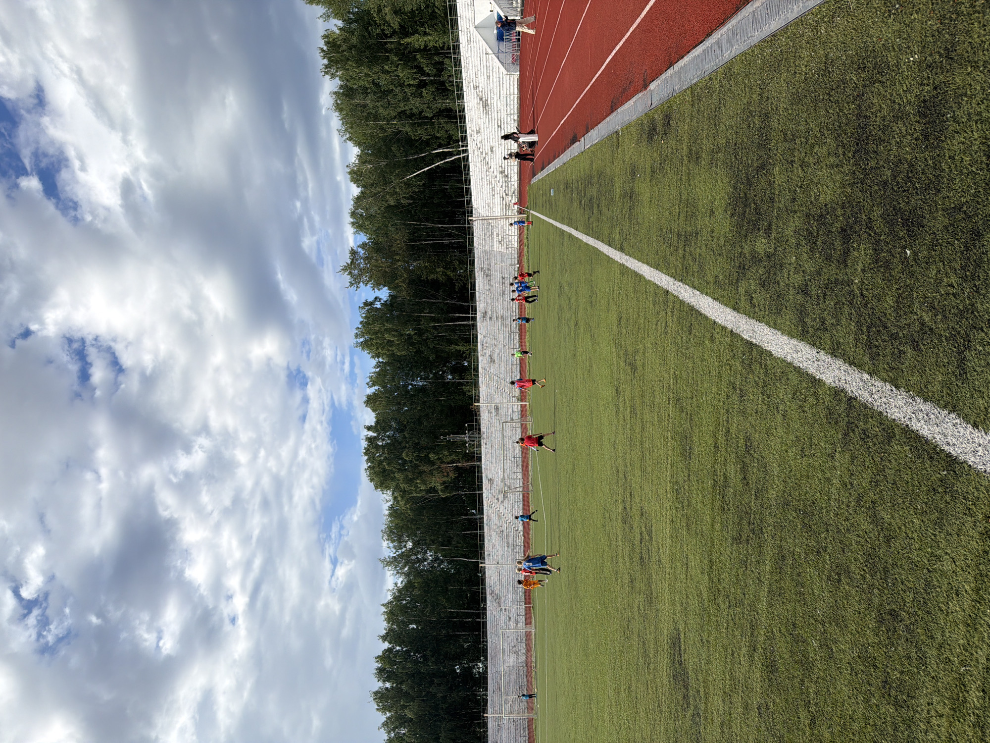 This image depicts a group of young people engaged in a sports activity on an outdoor artificial turf field. The field has a red running track bordering one side and is marked with white lines. Several individuals, likely athletes, are scattered across the green turf, some appearing to be in motion, possibly training or playing a game. In the background, there are bleachers and a dense line of trees, suggesting the location is a sports complex or stadium surrounded by nature. The sky above is filled with a mix of white and gray clouds, indicating a partly cloudy day, and the lighting suggests it is daytime. The overall scene conveys an atmosphere of active recreation. There is no visible text in the image.