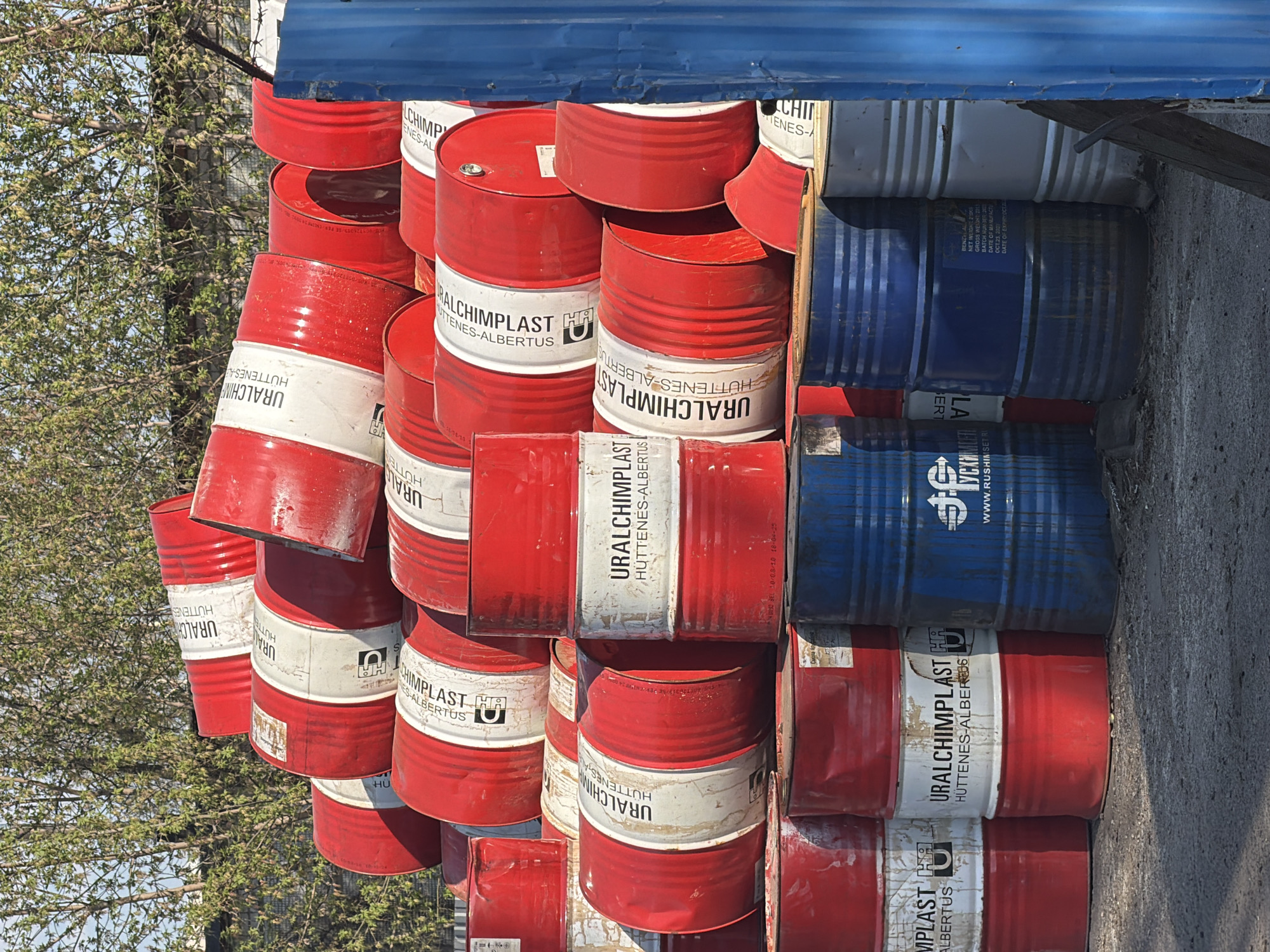 The image depicts a stack of cylindrical barrels primarily painted red, with some blue and white barrels interspersed. The barrels are labeled with "URALCHIMPLAST" and "HUTTENES-ALBERTUS." The barrels appear to be arranged haphazardly, some slightly tilted, creating a makeshift structure. To the left of the barrels, there is a tree with green foliage. Above the barrels, there is a blue metallic overhang. Based on the labels on the barrels and other information available in the images, the location is likely in or around the city of Chaikovsky, Russia.