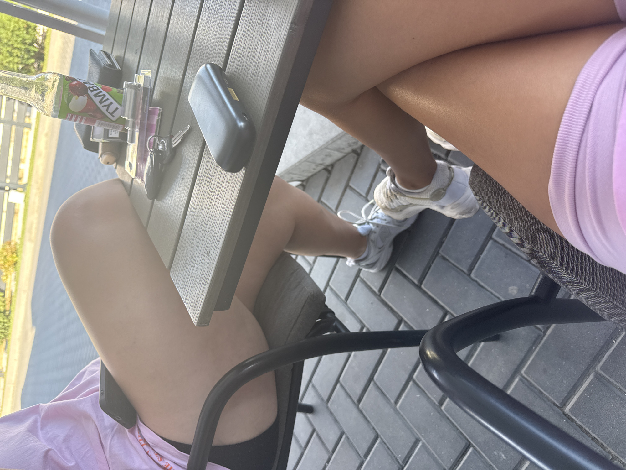 The image depicts two individuals seated at a table outdoors, presumably at a cafe or restaurant. The foreground shows a portion of one person's legs, clad in a light pink garment. Their right leg is crossed over the left, and they are wearing white sneakers. The background reveals another person's legs, wearing black shorts and a pink t-shirt, with a black smartphone resting on their lap.
On the table, several objects are visible, including a glass bottle with a green label that reads "TYMBA," a wallet, a cigarette lighter, keys, and a black electronic device. The table surface appears to be made of wood. The individuals are seated on chairs with dark metal frames and grey cushions. The ground beneath the table is paved with dark rectangular tiles. In the distance, a white fence and some greenery are discernible, suggesting an outdoor setting in a developed area. The lighting indicates it is daytime. The scene is likely located in the city of Comrat, Moldova, as specified in the prompt.
