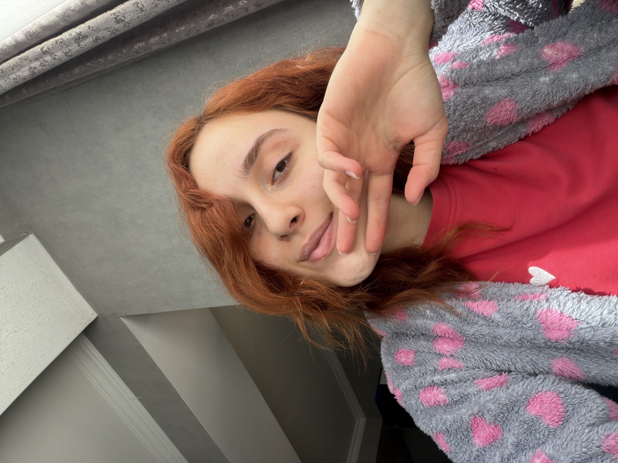 A person with reddish-brown hair is seen from a low angle. The person's face is slightly tilted, with their right hand raised in front of their face. The person is wearing a red shirt with a white heart shape on the chest and a gray fuzzy robe with pink polka dots. In the background, there are gray walls and a curtain. The location cannot be determined from the image.