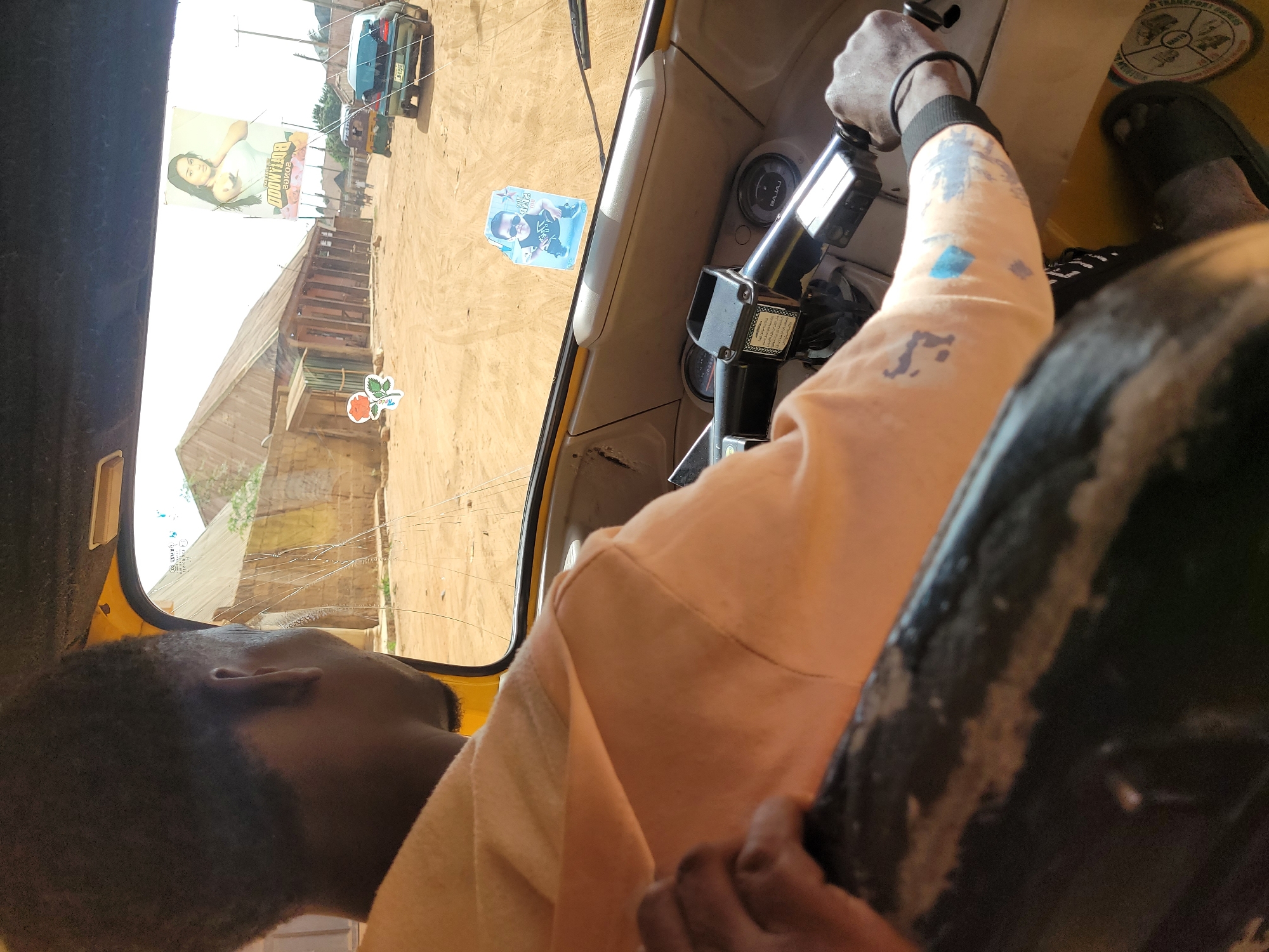 The image captures a first-person perspective from inside a Keke Napep (auto-rickshaw) in Jos, Nigeria, during daylight hours. The interior of the vehicle is visible, along with a glimpse of the street outside through the windshield and side window.
In the foreground, a person's head, facing left, is visible in the bottom left corner, suggesting they are a passenger or driver. To the right, another person's arm, clad in a peach or light orange long-sleeved top with blue and grey paint or tattoo marks, holds the vehicle's handlebar or steering mechanism. This person is wearing a black bracelet and black flip-flops are visible on their feet. The dashboard shows gauges and the "BAJAJ" brand logo, consistent with Keke Napep models. A circular sticker, partially visible near the driver's feet, has "Road Transport" text.
Outside the vehicle, a dusty, possibly unpaved road is visible, with several simple buildings and structures lining it. The sky appears bright, indicating clear weather. A large billboard with a woman's image and mirrored text, where "BOLLYWOOD" is clearly readable, stands prominently on the left. Further down the road, a dark-colored car is parked or driving. A small blue sticker featuring a man's face and mirrored text is affixed to the windshield. Another sticker of a red rose with the word "Rose" is visible on the outside of the windshield near a building. Notably, the windshield on the right side has a visible crack.