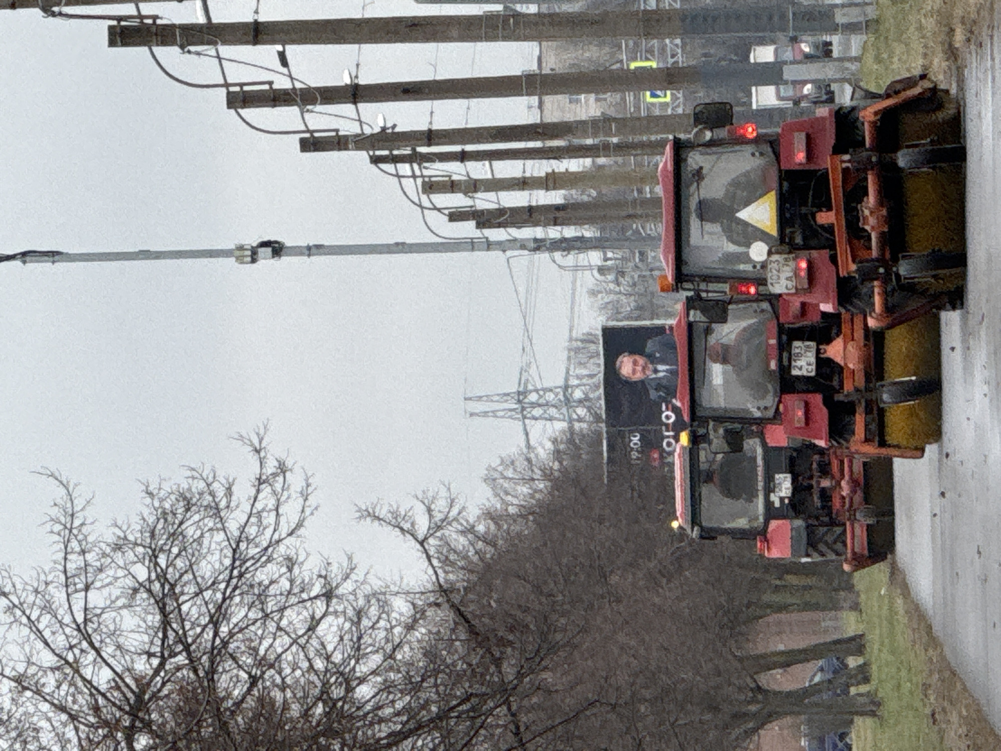 The image shows a street sweeper truck on a wet road in what appears to be Saint Petersburg, Russia. Bare trees are visible along the road. A billboard featuring a man in a suit is present. Power lines and utility poles run along the street. The sky is overcast.