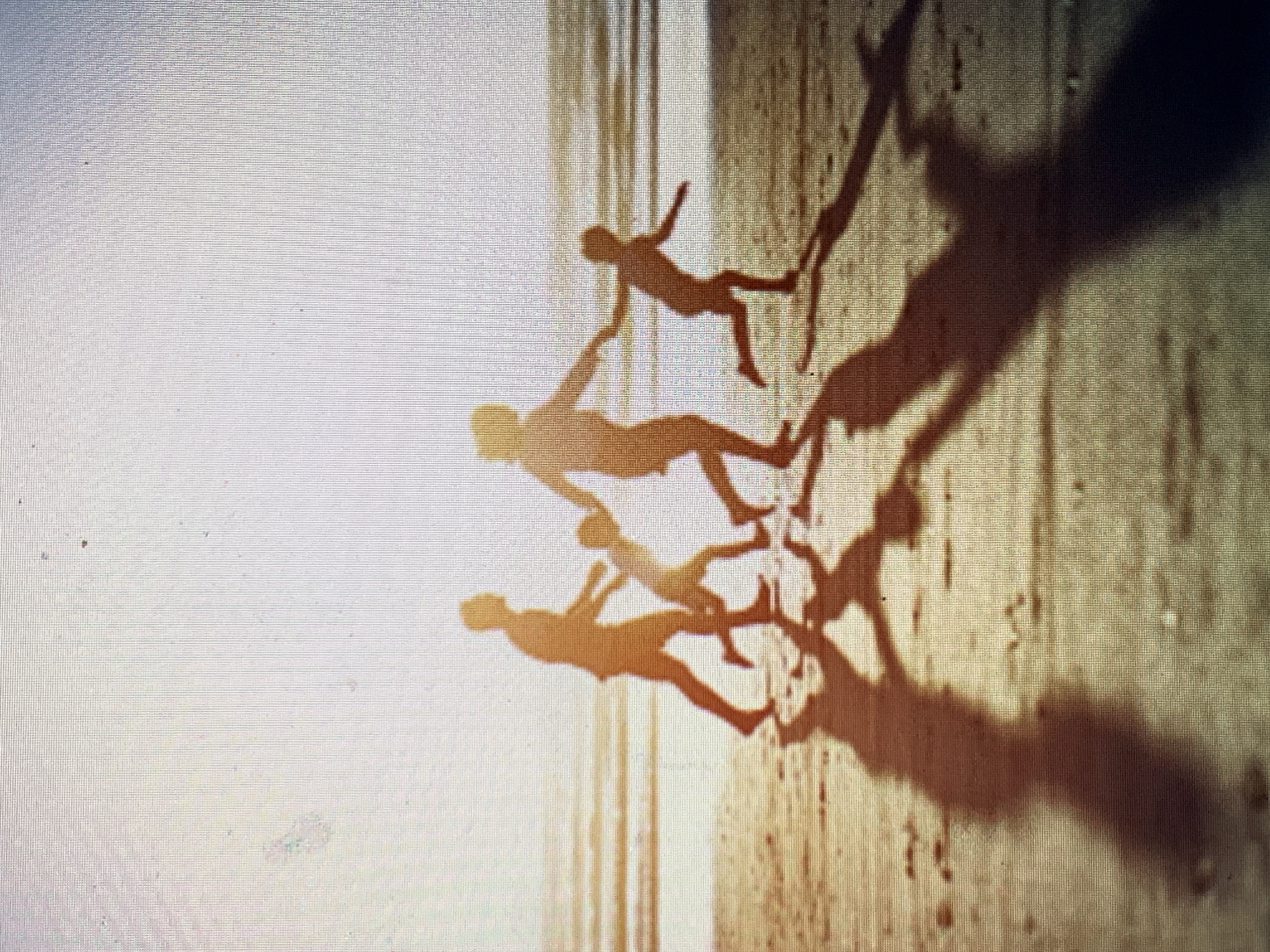 The image shows a beach scene with four figures casting long shadows. The figures appear to be holding hands in a chain, and their shadows are elongated due to a low angle of sunlight. The beach has a wet, reflective surface, suggesting it's near the water's edge. The environment suggests a coastal setting with sandy terrain. The image does not show details that allow for location verification within Barcelona, Spain.