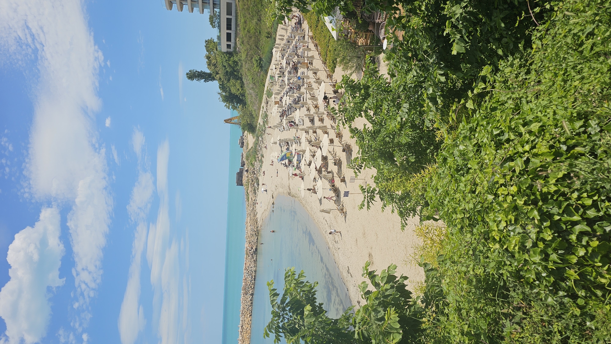 The image shows a beach scene at Kabakum Beach in Varna, Bulgaria. The beach is crowded with people, mostly sunbathing on lounge chairs under white umbrellas. The beach is sandy and slopes down to a calm, clear body of water. A rock barrier runs along the left side of the water. There is a high-rise building along the coastline in the background. A crane is visible on the shoreline near the building. Lush greenery is visible along the upper right side of the frame. The sky is blue with scattered white clouds.