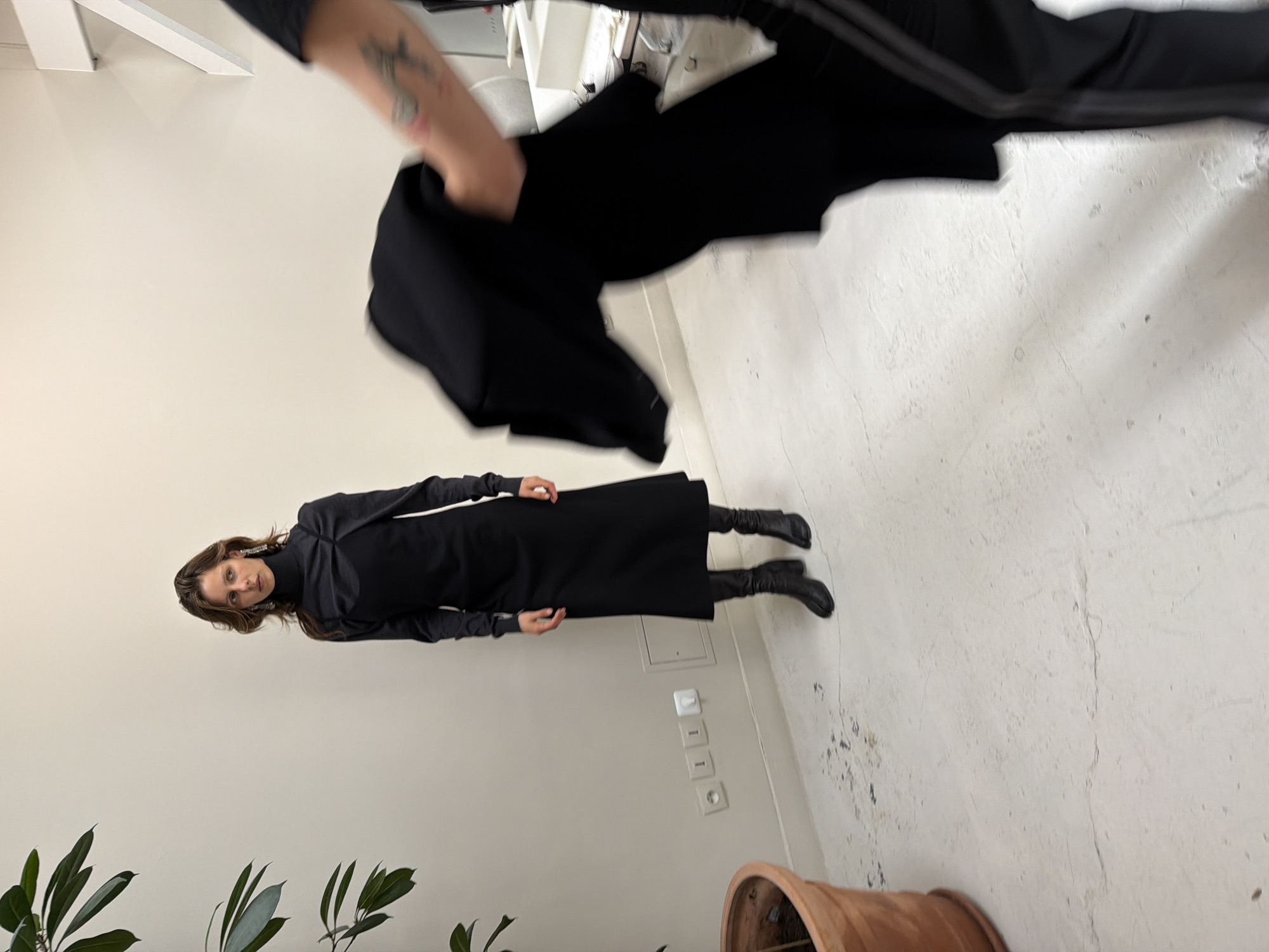 A full-length portrait of a woman with long, light brown hair wearing a long, black dress with long sleeves and black boots. She stands against a plain white wall near a potted plant with green foliage. Electrical outlets are visible along the wall. A hand holding a black garment is present in the upper portion of the image. The background is blurred, suggesting the shot was taken indoors, likely in an atelier or studio setting. The location cannot be determined to be in Paris.