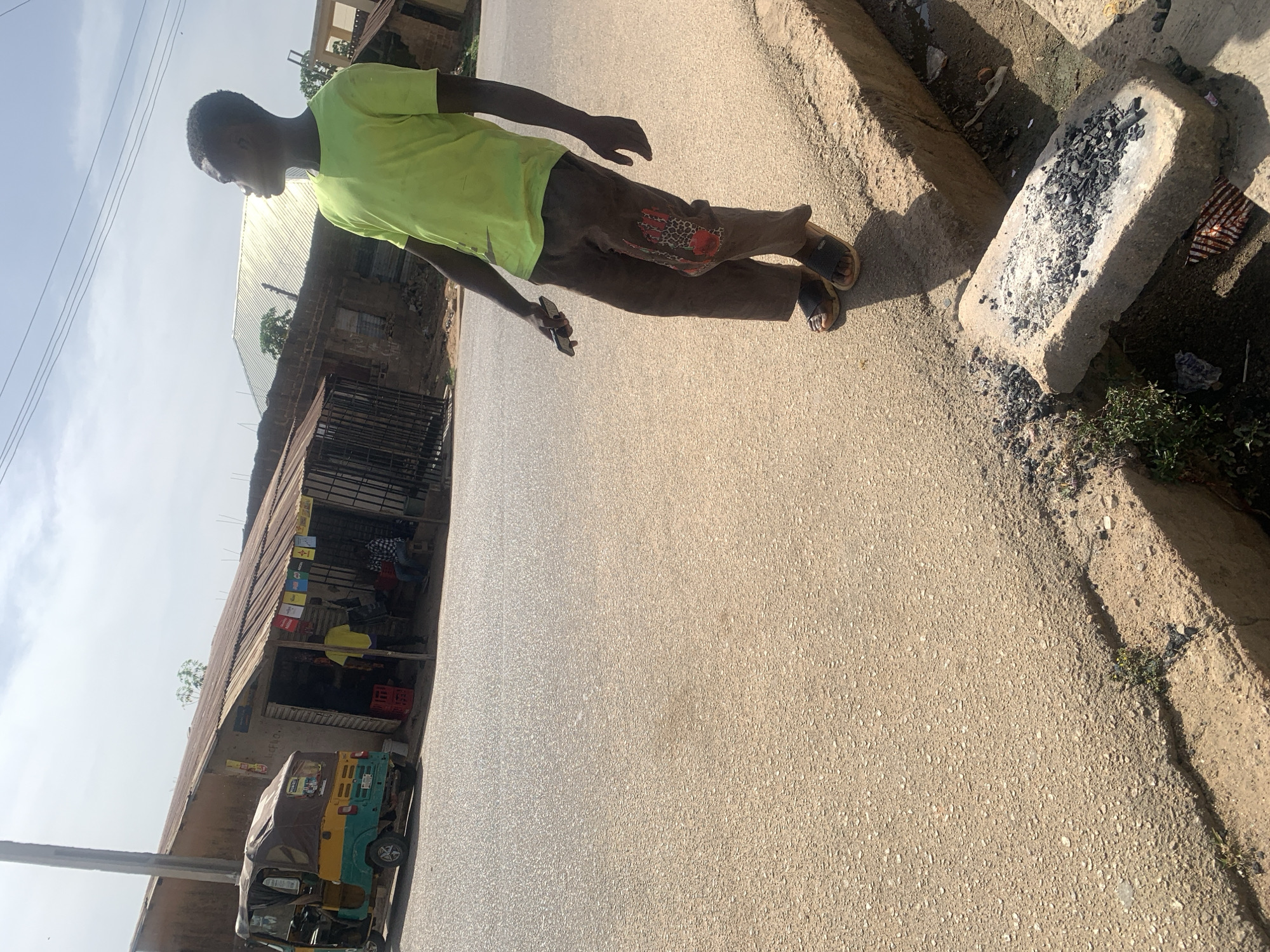 A mid-day shot shows a street in Jos, Nigeria. A young man in a light green t-shirt and brown pants with a decorative patch is walking on the road while looking at a phone in his hand. He is wearing sandals. To his left, a shop is visible, with people sitting inside. A "keke", a type of auto rickshaw common in Nigeria, is parked in front of the shop. To the right of the man, a rectangular cement block filled with what appears to be burnt charcoal sits on the edge of the paved road, next to a dirt shoulder with sparse vegetation. The sky is partly cloudy.