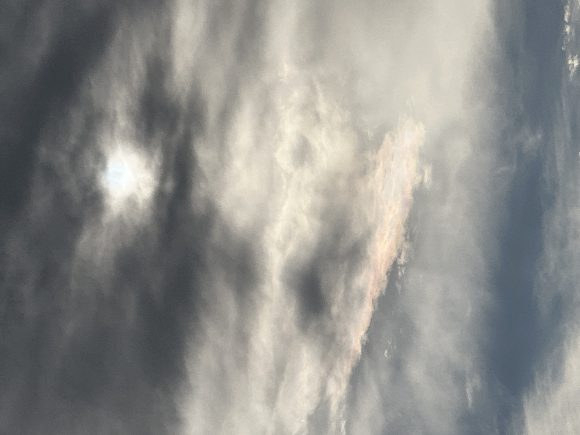 The image shows a daytime sky filled with layered clouds. The clouds range in color from dark gray to light gray and white. A bright spot, possibly the sun, is partially obscured by the clouds. A small section of the clouds exhibits iridescent coloration, displaying a range of pastel colors. The location of the image is Zelenikovo, Bulgaria. There are no people or other specific objects in the frame. The main subject is the sky and clouds.