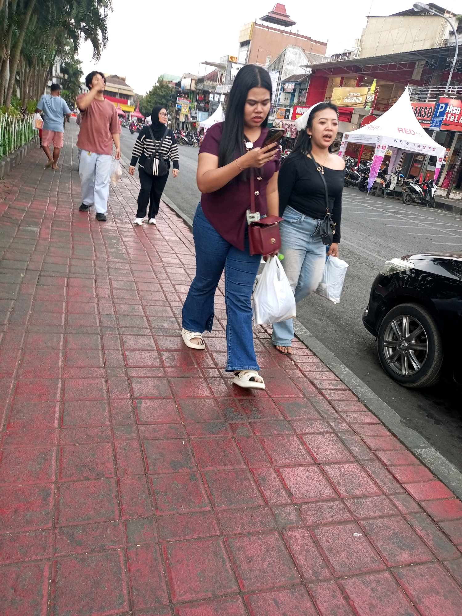 In Batu, Indonesia, a group of people are walking along a sidewalk with red paving stones. In the foreground, two women are walking towards the viewer. The woman on the left, wearing a maroon shirt and blue jeans, is looking at her phone and carrying a red purse. The woman on the right, wearing a black off-the-shoulder top and blue jeans, is looking forward and carrying two white plastic bags. Behind them, a man in a brown t-shirt and light blue pants is walking away from the viewer, holding a plastic bag. Further down the sidewalk, a man in a blue t-shirt and pink shorts is also walking away from the viewer. In the background, there are shops and tents, suggesting a marketplace or event. A black car is parked on the street to the right of the sidewalk. The sky is overcast.