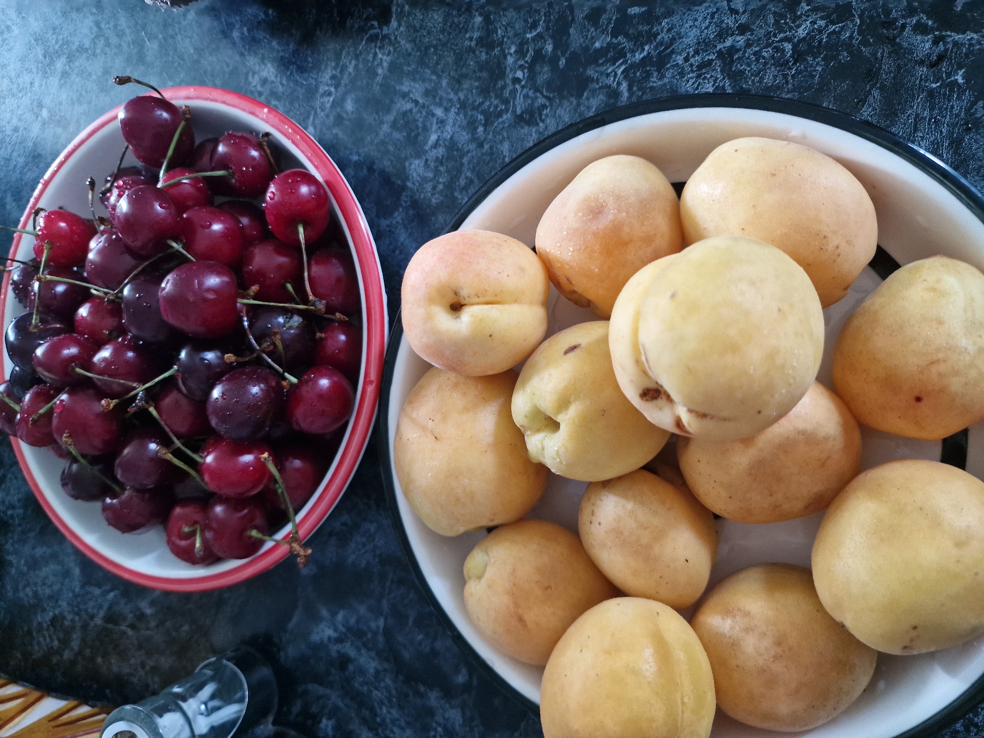 Two white bowls filled with fruit sit on a dark, textured countertop. The upper bowl, slightly smaller and with a red rim, contains a pile of dark red cherries. The cherries are wet, suggesting they have been washed. The lower bowl, larger and with a black rim, is filled with yellow-orange apricots. Some of the apricots have slight reddish blush marks. The background countertop is a mottled dark blue and black. To the left of the bowls, a glass salt or pepper shaker with a silver lid is partially visible. The scene appears to be an indoor setting. The location of Kursavka, Russia is not visually evident from the image.