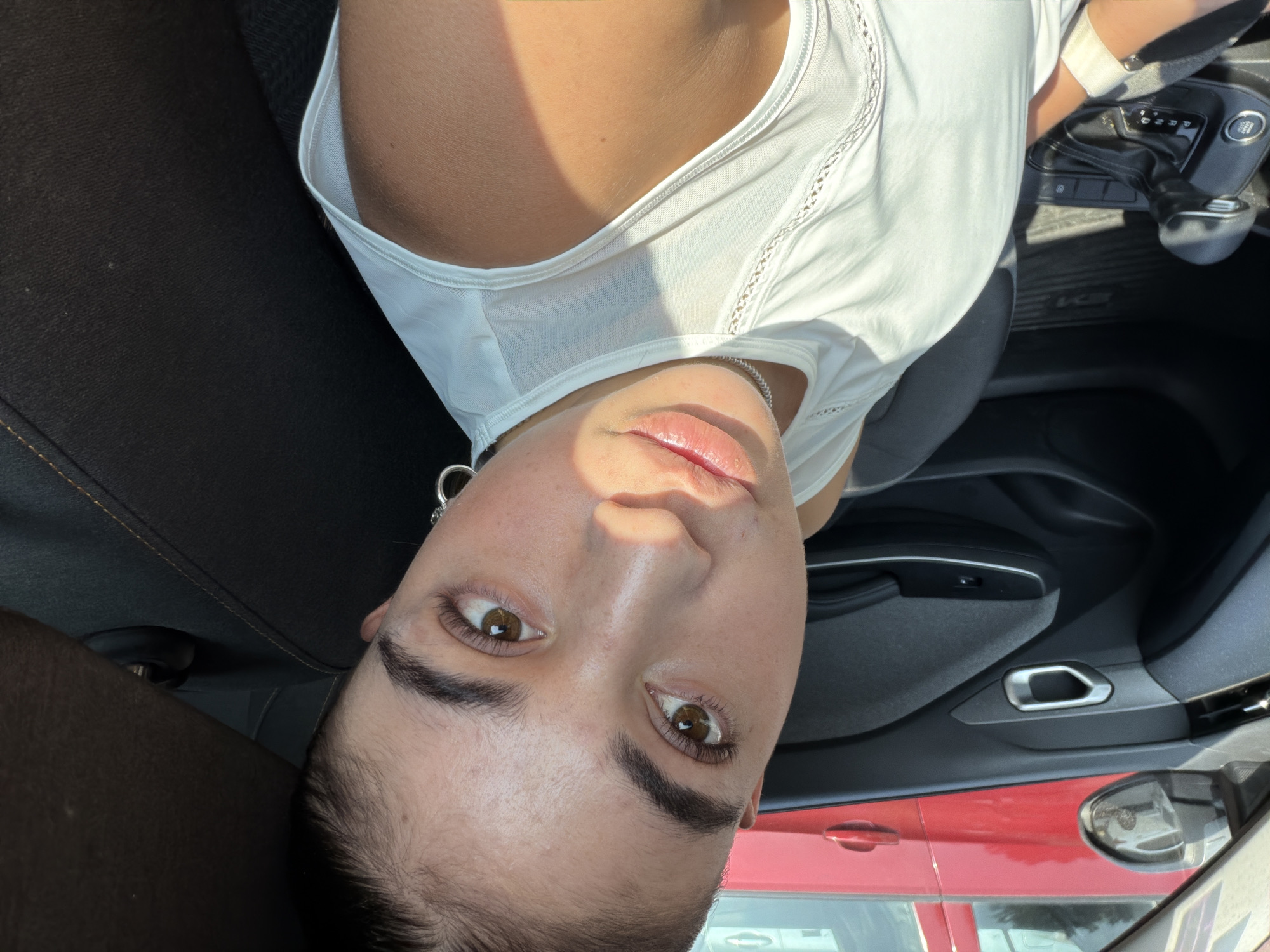 A woman is shown in a head-and-shoulders self-portrait taken from within a car. She is wearing a white top and a silver necklace. The car's interior is visible, with a gear shift and a door handle partially in view. Outside the car, a red vehicle can be seen through the window. The location is unidentifiable as Puebla, Mexico, based solely on the visual elements within the image.