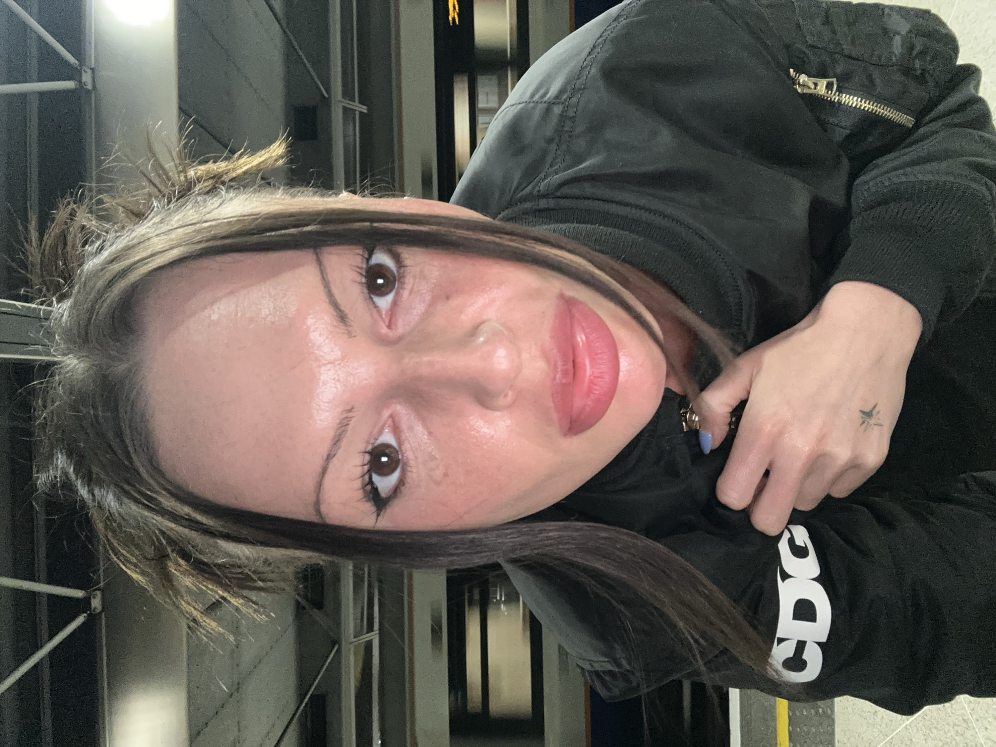 A medium close-up portrait of a woman indoors. The woman has long, brown hair with blonde highlights, brown eyes, and fair skin. She is wearing makeup, including eyeliner and lipstick. She is dressed in a black bomber jacket with a visible gold zipper. The word "CDC" in white block letters is partially visible on the sleeve of the jacket. Her left hand is visible, adorned with a small tattoo on the wrist. In the background, vertical metal bars are seen, suggesting an urban setting. Based on visual elements and available information, a possible location in the city of London, United Kingdom is a train station due to the metal vertical bars and door like structures in the background.