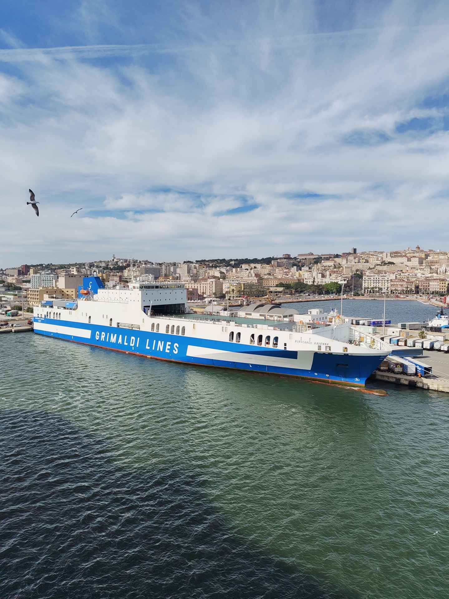 A large Grimaldi Lines cargo ship, named "Eurocargo Ravenna," is docked in the port of Cagliari, Italy. The ship is predominantly white with a blue stripe and blue accents. The cityscape of Cagliari forms the backdrop, with buildings stretching across the horizon. The sky is blue with scattered white clouds and contrails. In the foreground, dark water is visible, with two birds (possibly seagulls) flying in the upper left quadrant. The ship is docked at a pier with shipping containers and vehicles visible nearby.
