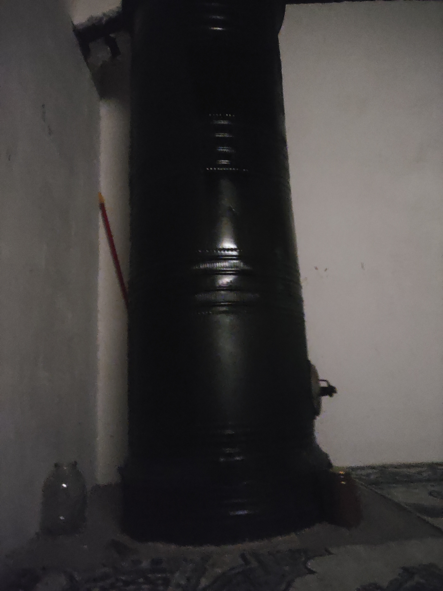 The image depicts a dark, tall, cylindrical object, possibly a stovepipe or a storage tank, positioned vertically in the center of the frame. It is a matte black color with horizontal ribbed sections visible along its surface. To the left of this object, a white wall forms a corner with another wall that appears to be a darker gray. Leaning against the white wall is a thin, red object, potentially a broom or a stick. In the bottom left corner, a clear glass jar with a rounded base is visible. The floor is covered with a patterned material, suggesting it may be a rug or tiled surface. The lighting is dim, casting shadows and obscuring details in the background. The overall scene appears to be indoors, possibly in a storage area or a room with a heating apparatus. The location in Kiyatabad, Uzbekistan, is not visually evident from the provided image.