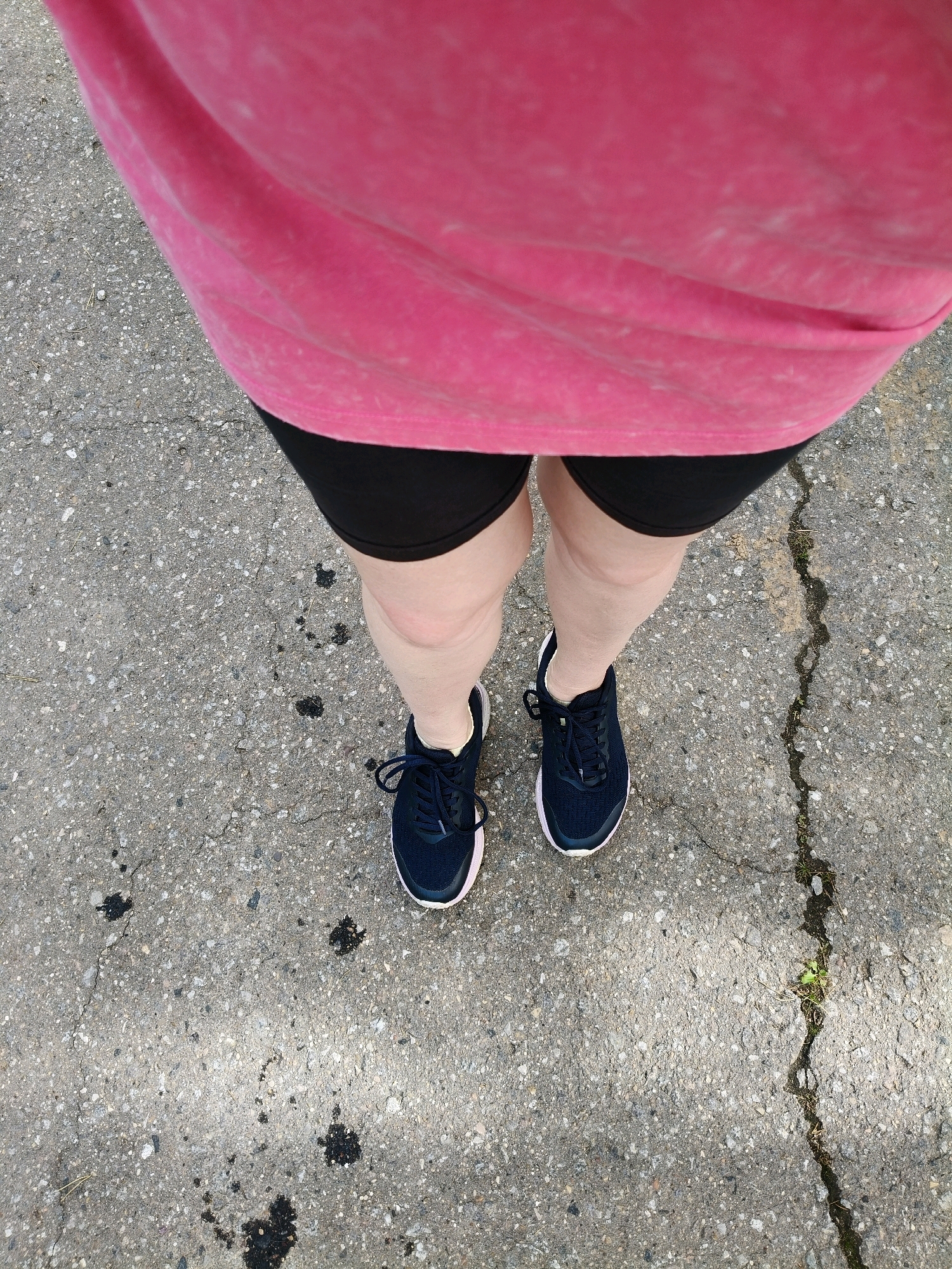 A person stands on a cracked asphalt surface. They are wearing a bright pink t-shirt, black shorts, and dark blue athletic shoes with white soles and pink accents. The perspective is from above, looking down at the person's legs and feet. The asphalt has scattered dark spots, possibly oil or tar, and a prominent crack runs vertically down the right side of the frame. A small amount of green vegetation is visible growing from the crack. The lighting suggests an outdoor, possibly daytime setting, with some dappled shadows on the ground.