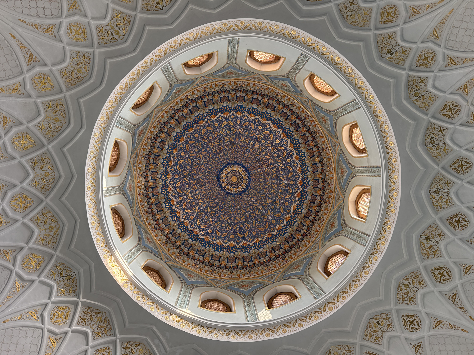 The image captures an upward view of the dome and ceiling of a structure, likely a mosque, in Tashkent, Uzbekistan. The dome is centrally positioned, showcasing intricate mosaic patterns in blue and gold radiating outward from the apex. A ring of small windows encircles the base of the dome, allowing light to filter in. The surrounding ceiling is adorned with geometric patterns and floral motifs in white and gold. No people are visible in the shot. The scene depicts the interior architecture of a religious building, emphasizing the ornate decorations.