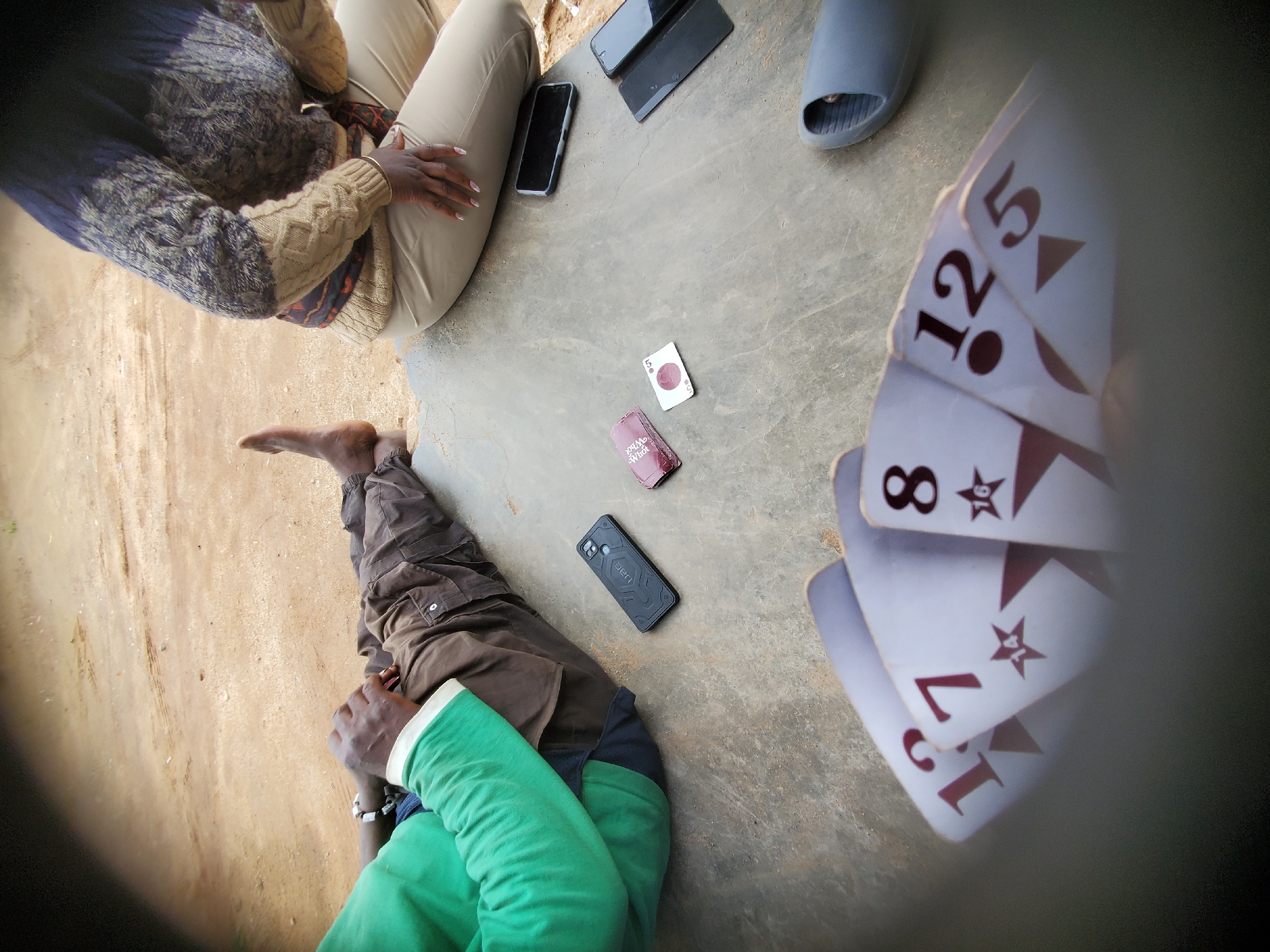 The image shows a group of people on a concrete surface, possibly outdoors. In the foreground, a person is lying on their side with their legs bent. They are wearing a green long-sleeved shirt and brown cargo pants. Their hands are visible, one resting on their stomach.
Another person is seated to the left, wearing a patterned sweater and light-colored pants. Their hand is resting on their thigh, and they are wearing a gold bracelet.
Scattered on the ground are several objects, including mobile phones, a pair of blue slides, and playing cards. Some of the playing cards are spread out, revealing numbers such as 8, 12, 5, and 7, along with star symbols. One card has text that reads "You Me What". Two mobile phones are visible, one black and one dark gray, positioned near the seated person. Another black mobile phone is closer to the person lying down. A blue slide is visible on the right side of the image. The ground appears to be a mix of concrete and dirt.
The lighting suggests it could be daytime, with natural light illuminating the scene. The overall mood appears casual, suggesting a game or a gathering. The location cues, such as the concrete and dirt, are consistent with an outdoor setting in Bukuru, Nigeria.
