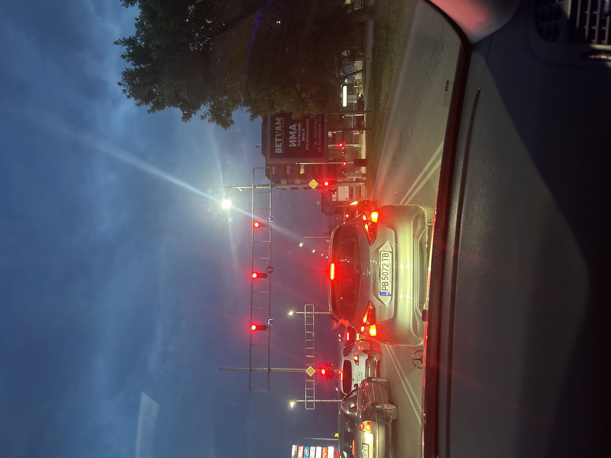 The image depicts an urban street scene at dusk or early night in Plovdiv, Bulgaria, observed from the interior of a stationary vehicle. The foreground reveals the dark dashboard of the vehicle, distinctly featuring a metallic Mercedes-Benz emblem.
Directly ahead, the road is occupied by multiple vehicles queuing at a red traffic light. A silver Nissan Pulsar, displaying Bulgarian license plate "PB 5072 TB" (PB denoting Plovdiv), is prominently centered with its red brake lights illuminated. To its left, a dark-colored sedan with Bulgarian license plate "PB 5087 KT" is also stationary. Further ahead in the queue, a white SUV/hatchback with license plate "PB 1050 MA" is visible. All visible vehicles have their brake lights active, signaling a stop.
Overhead, a gantry structure spans the road, holding several red circular traffic lights for multiple lanes, alongside illuminated white arrow signs indicating left turn and straight-ahead directions. Additional red traffic lights are mounted on poles at a lower level. Bright streetlights line the road, casting strong illumination and creating lens flares.
On the left side of the street in the background, commercial buildings are partially visible, featuring fragmented signs for "MALL PARK," "Techno," and what appears to be part of an "IKEA" logo. On the right side of the street, a multi-story building features a large billboard prominently displaying "BETVAM" in Latin script and "ИМА ПЪТУВАНИЯ ИМА И ПРЕЖИВЯВАНИЯ" in Cyrillic, with smaller Cyrillic text below referencing "18+ ХАЗАРТЪТ Е ОТ РАЗВИВАНЕ НА..." (translating to "THERE ARE TRAVELS THERE ARE AND EXPERIENCES" and "18+ GAMBLING IS FROM THE DEVELOPMENT OF..."), confirming the Bulgarian context. A large, dense-leafed tree stands to the right of this building. The sky above is a dark blue, overcast with clouds, typical of evening conditions. No specific individuals are discernible in the scene.