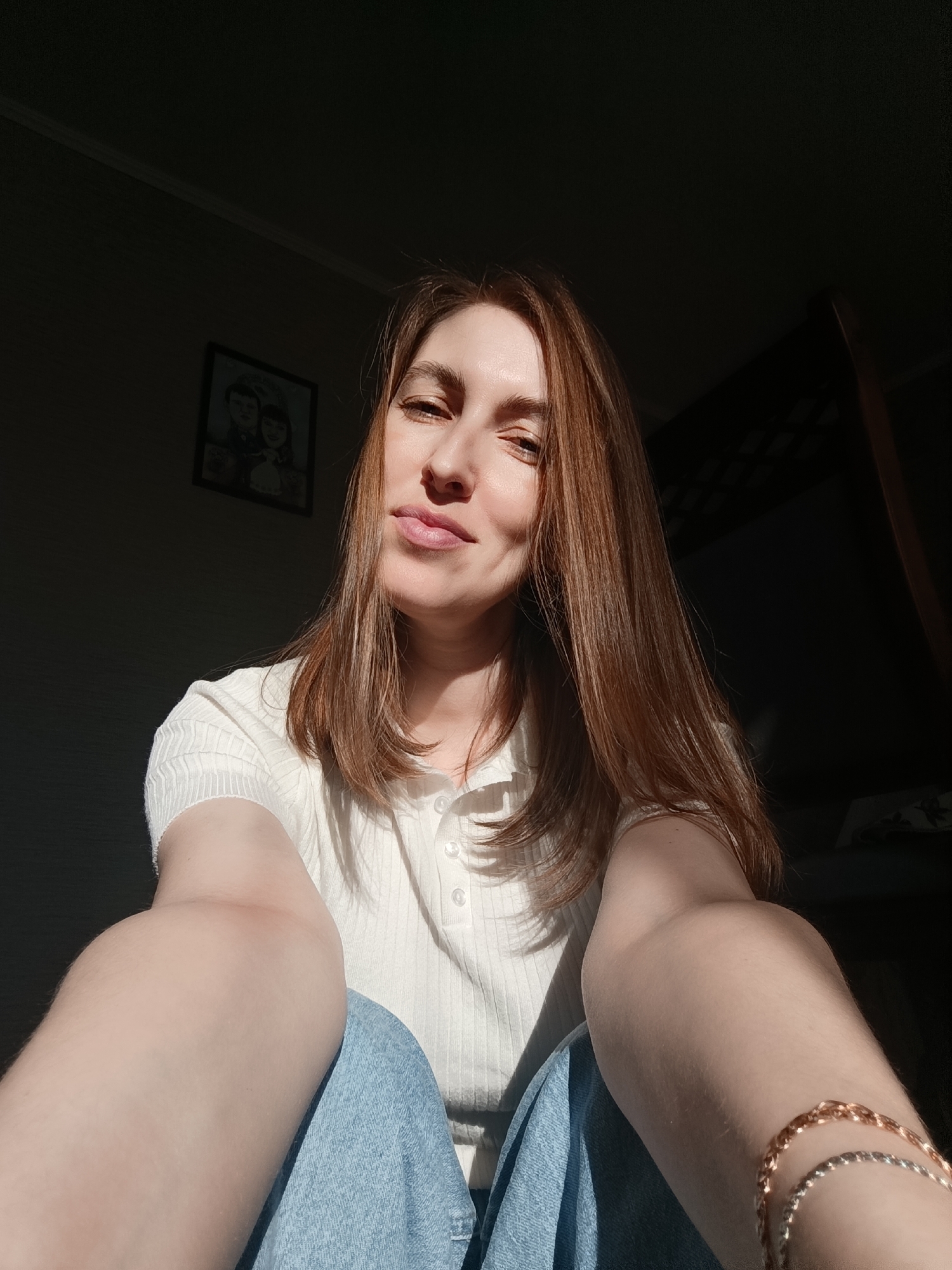 A young woman with auburn hair sits facing the camera in a dimly lit room. She wears a white, short-sleeved, button-down shirt and blue jeans. Two bracelets are visible on her right wrist. A picture hangs on the wall behind her. A partial bunkbed frame is visible in the background. The location is in the city of Фоки, Russia.