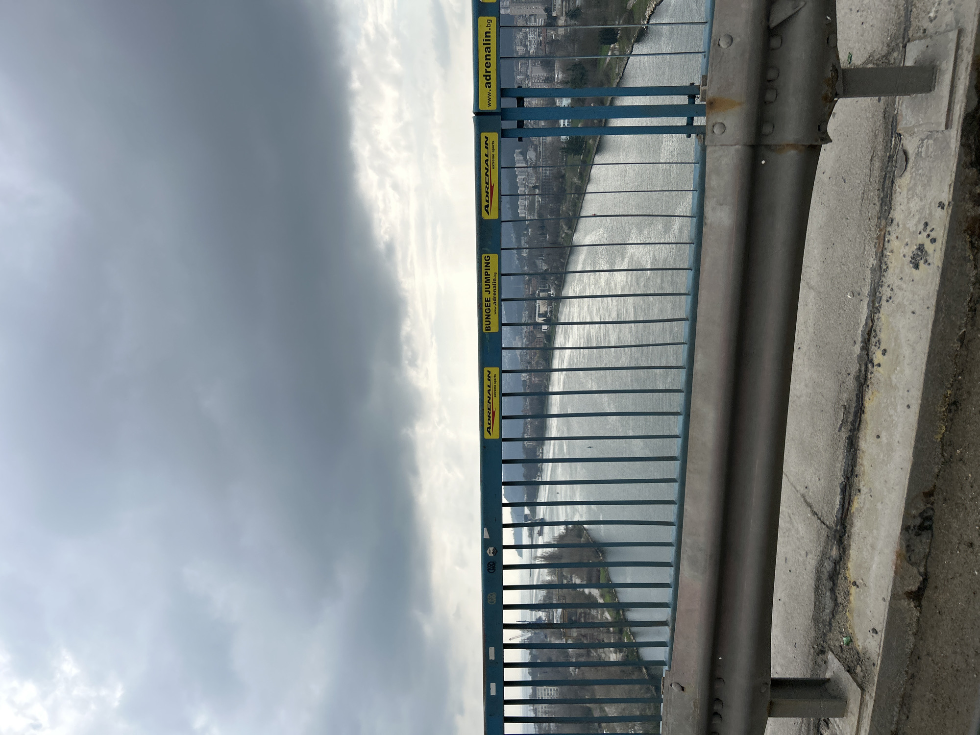 The image shows a view from a bridge, likely overlooking a river in Unknown, Unknown. A blue metal fence with yellow advertisements for "Adrenalin extreme sports" lines the side of the bridge. Beyond the fence, there's a body of water reflecting sunlight. A city skyline is visible in the background. The sky is overcast with gray and white clouds. The foreground includes a concrete barrier and a metal guardrail adjacent to the fence.