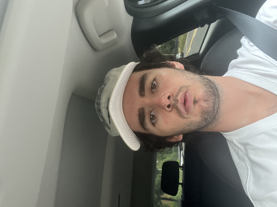 A young adult male is depicted from the chest up, seated in the passenger side of a vehicle in Unknown, Unknown. He has dark, wavy hair, a short dark beard, and brown eyes directed towards the camera with a neutral expression, mouth slightly open. He is wearing a white t-shirt and a light-colored baseball cap with dark embroidery. A black seatbelt is secured across his right shoulder and chest. The vehicle's interior includes a light-colored headliner and a black headrest of a rear seat to the subject's left. Through the vehicle's windows, a paved road lined with green foliage is visible under ambient daylight, with a light-colored vehicle in the distance.