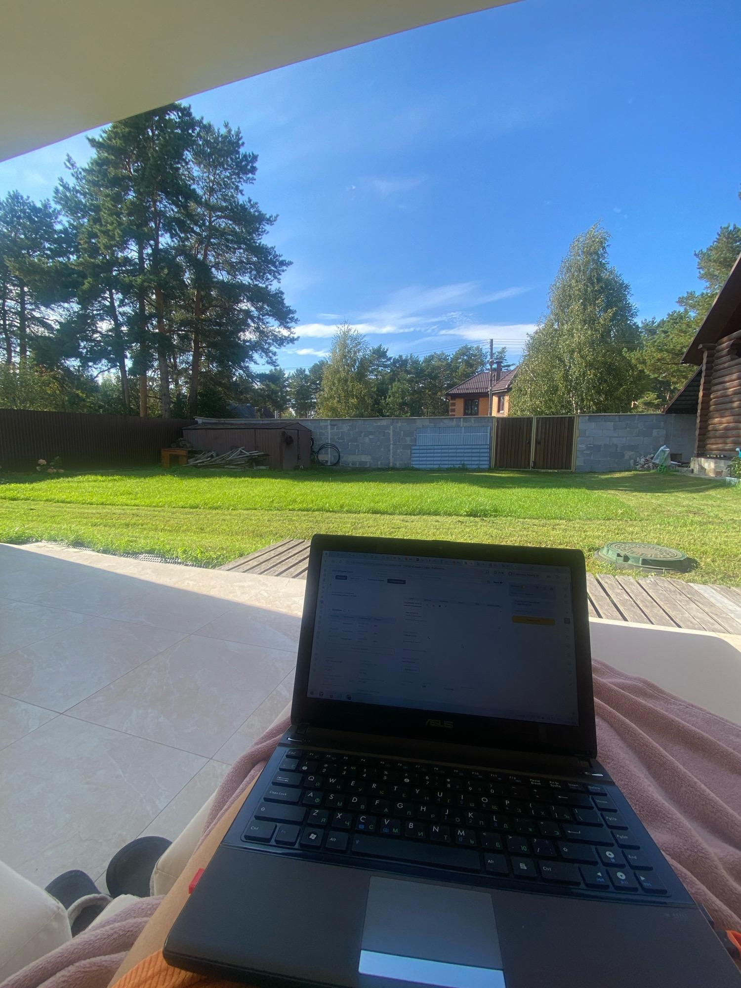 The image shows a personal laptop on a lap, with a blurred background of a backyard. The laptop is open and appears to be displaying a spreadsheet or data entry form. The background shows a green lawn, trees, a wooden fence, and a brick wall. The sky is blue with some scattered clouds. The image appears to be taken from a seated position, looking slightly downwards at the laptop. There is no indication of the city of Perm, Russia, or any specific location.
