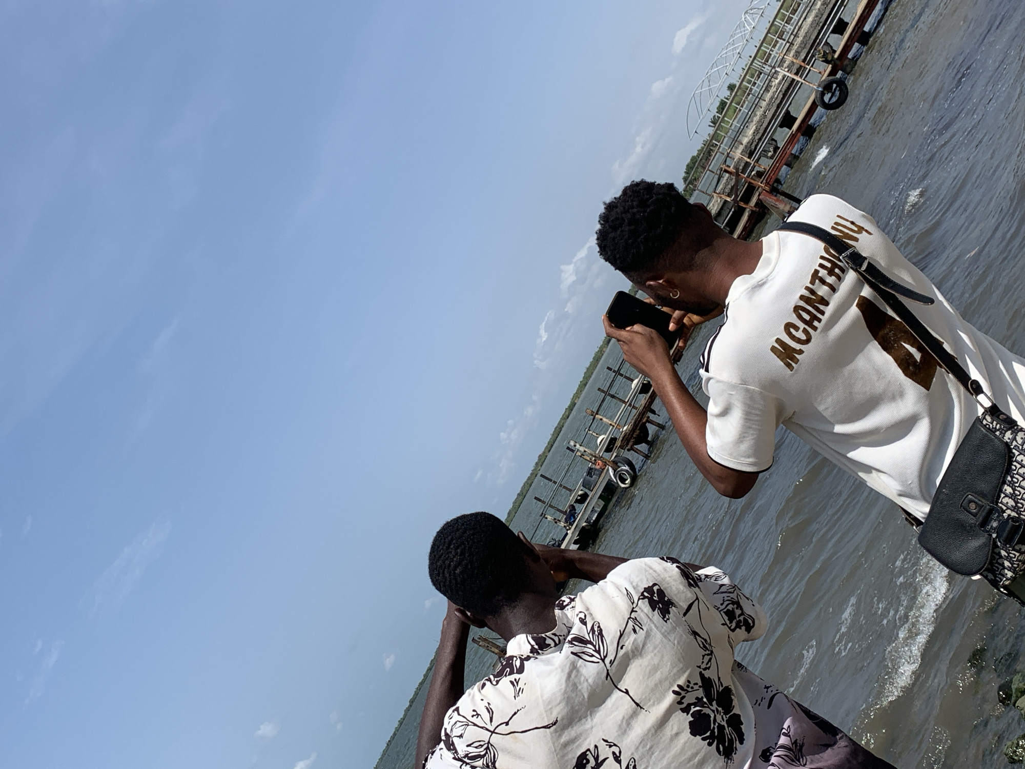 The photograph shows two men standing near a body of water, possibly a lagoon or river, in Ikorodu, Nigeria. One man, seen from the back, wears a white t-shirt with the name "MCANTHONY" printed on it. He holds a smartphone, possibly taking a photograph. A black shoulder bag is slung across his body. The second man, also seen from the back, wears a white shirt with a floral pattern and his head is in his hands. In the background, a dilapidated metal structure, possibly an abandoned dock or platform, extends into the water. The sky is light blue with some clouds. The water appears to be slightly choppy.
