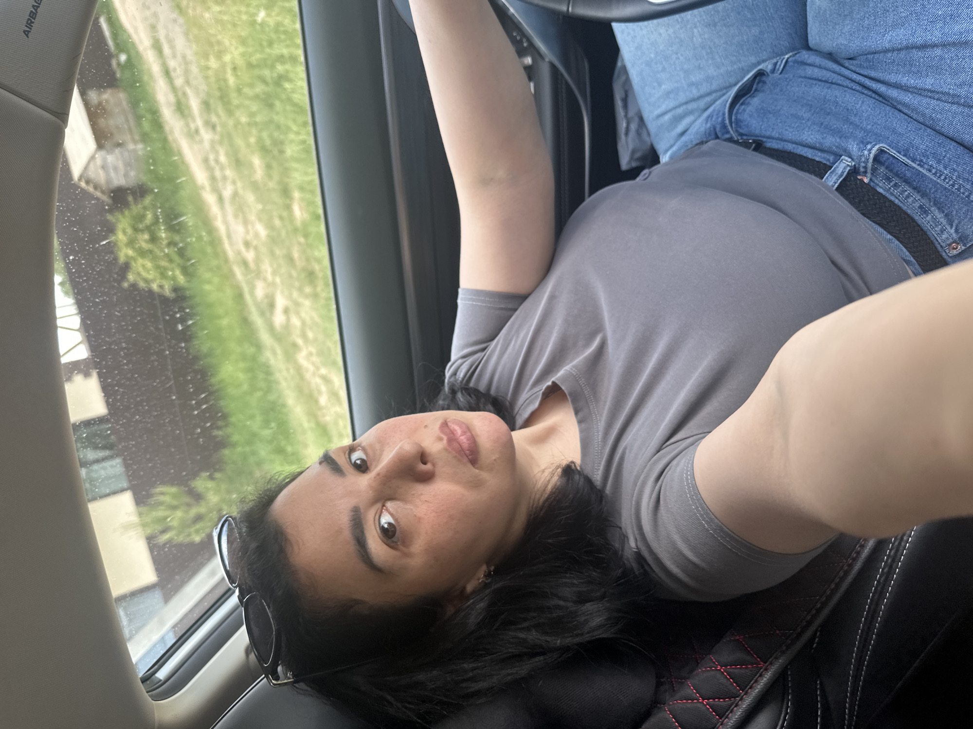 The image shows a woman lying in a car seat, holding the steering wheel. She is wearing a gray t-shirt and blue jeans with a black belt. Sunglasses are placed next to her on the seat. Outside the car window, a blurry landscape of green grass and buildings is visible. The location is unidentified.