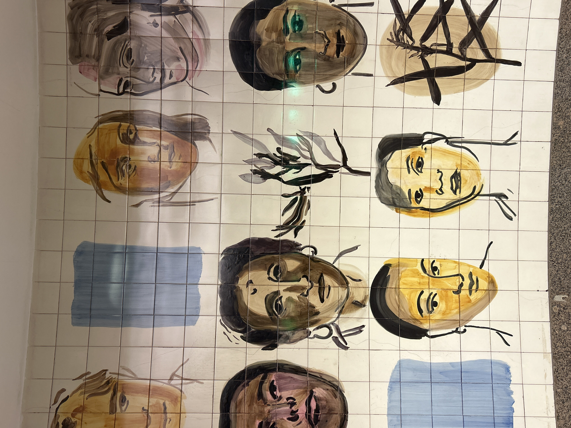 The image depicts a wall decorated with painted tiles. The tiles are arranged in a grid pattern and feature a series of stylized portraits of human faces in various skin tones, along with abstract patterns and a branch-like design. The faces appear to be painted in a naive or folk art style, with bold lines and simple shading. The color palette consists of earthy tones, including browns, yellows, blues and grays. The location is not identified in the image.