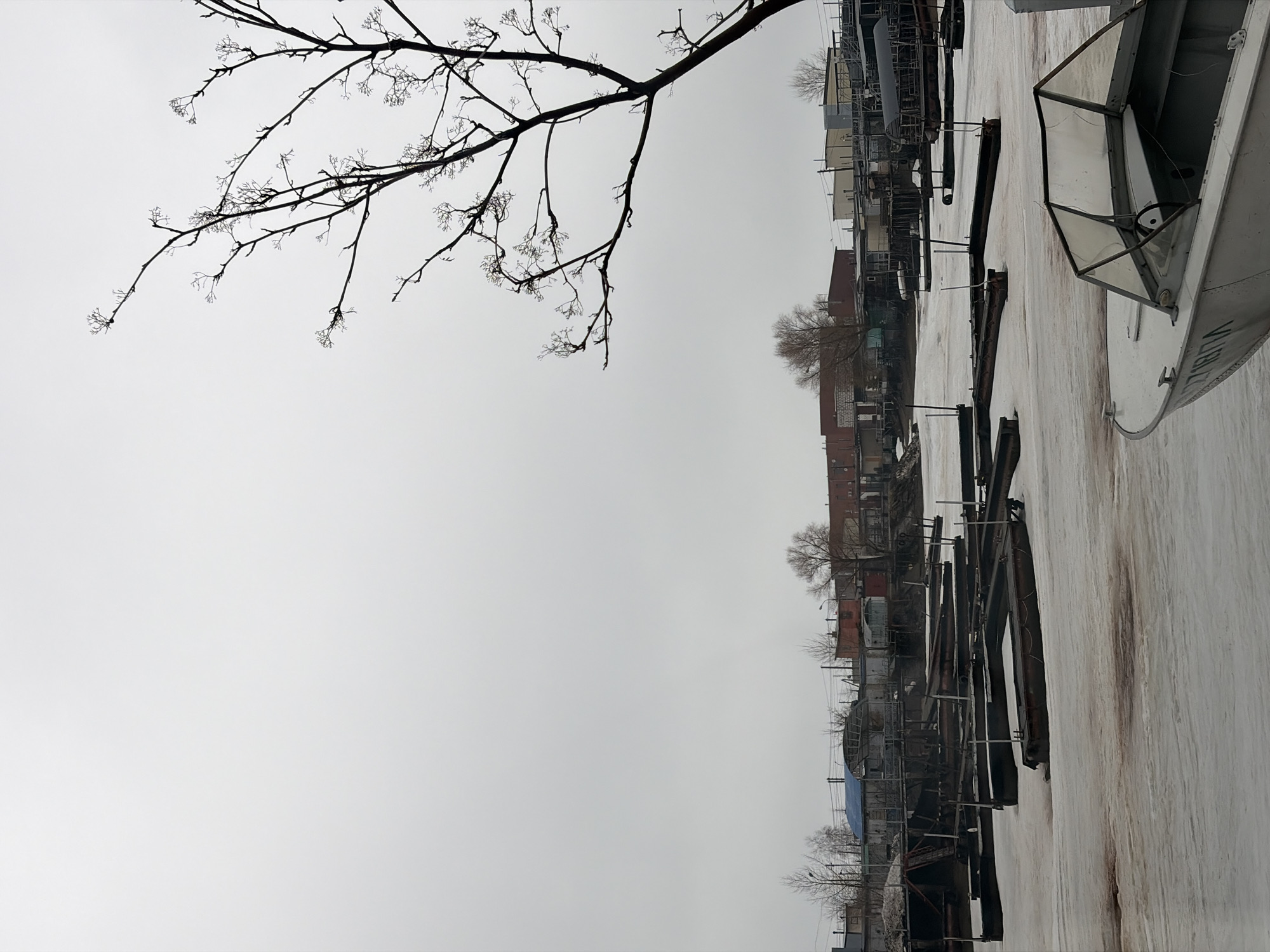 The image depicts a waterfront scene in Chaikovsky, Russia. The sky is overcast. A bare tree branch extends into the top left portion of the frame. Along the waterline, there are several boats, docks, and buildings. The docks are constructed of metal and wood. The water is partially frozen or covered in slush. A small, light-colored boat is in the foreground.