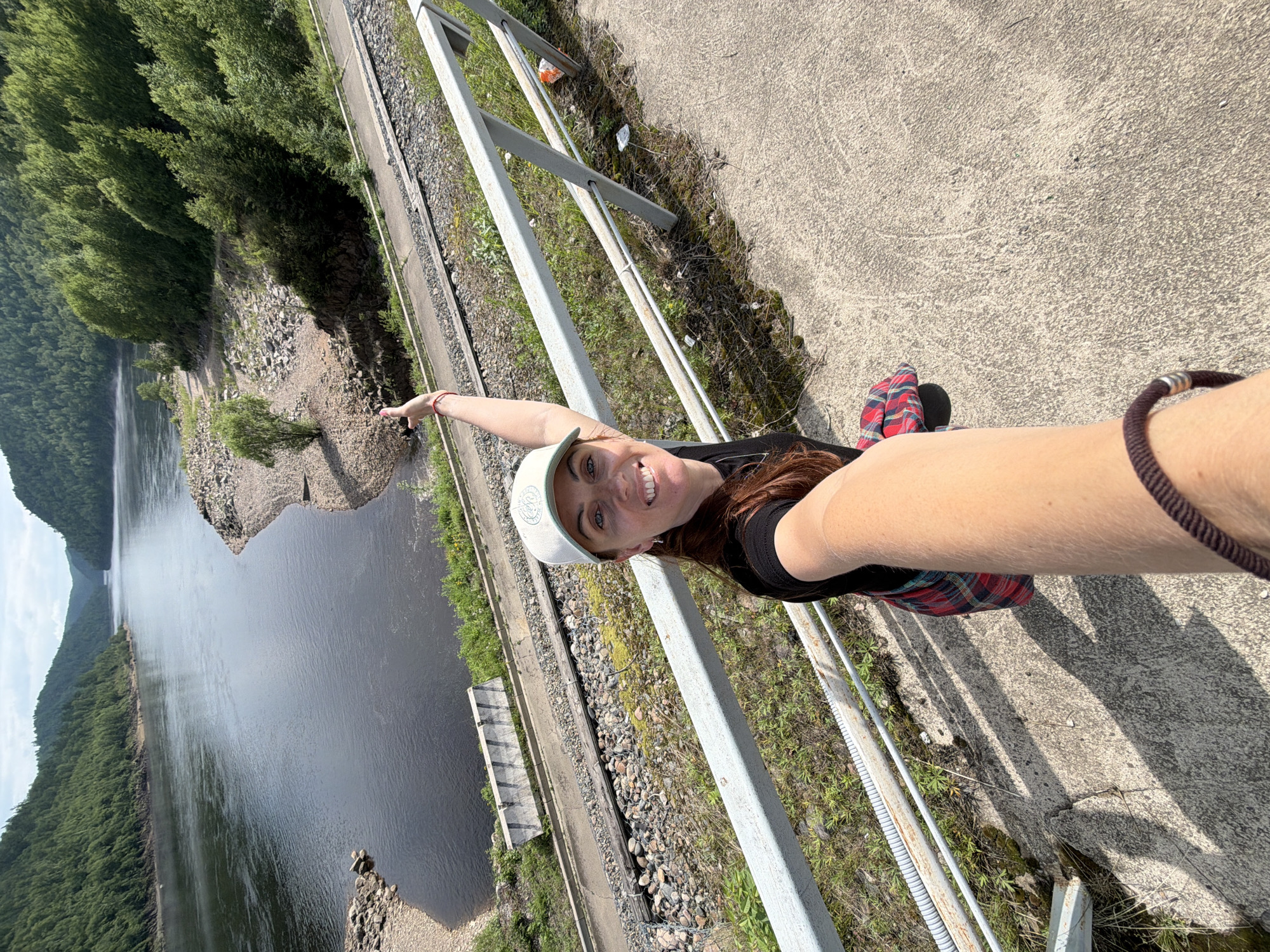 In Talakan, Russia, a woman with long, brown hair wears a black t-shirt, a red and black plaid skirt, and a light-colored baseball cap. She is taking a selfie with her arm extended, and she is smiling at the camera. Her left hand is raised, pointing towards the scene. She is standing on a concrete walkway next to a metal railing. In the background, a wide, dark river flows between steep, tree-covered hills. A set of train tracks runs alongside the river. The environment appears to be a scenic, natural area with a man-made element of a railway.