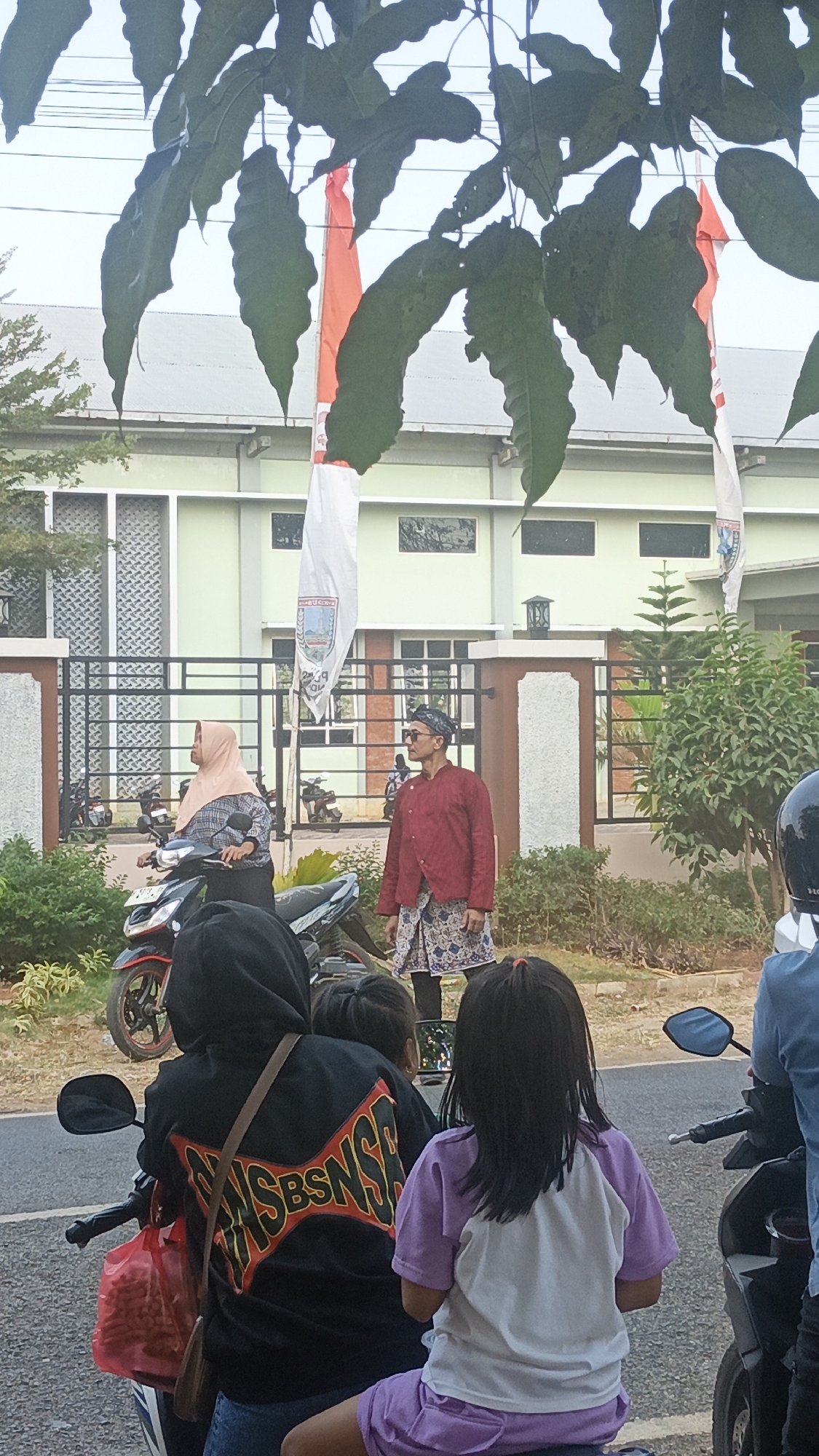 The image shows an outdoor scene with several people and vehicles. In the foreground, three individuals are seated on a motorcycle, with their backs to the camera. The person in the middle wears a black hoodie with a red and yellow graphic design on the back. To their right, a young girl wearing a purple t-shirt and shorts is visible. The person on the left, also on the motorcycle, is partially obscured.
Behind the motorcycle, a man stands facing left, wearing a red long-sleeved shirt and a patterned skirt over black leggings. He has a dark head covering and sunglasses. To his left, a woman is seated on another motorcycle, wearing a light pink hijab and a checkered shirt.
In the background, a light green building with multiple windows is visible. Two Indonesian flags, red and white, are flying from flagpoles in front of the building, partially obscured by green foliage in the upper portion of the image. The scene appears to be a street in front of a building, with parked motorcycles and greenery present. The lighting suggests it is daytime.