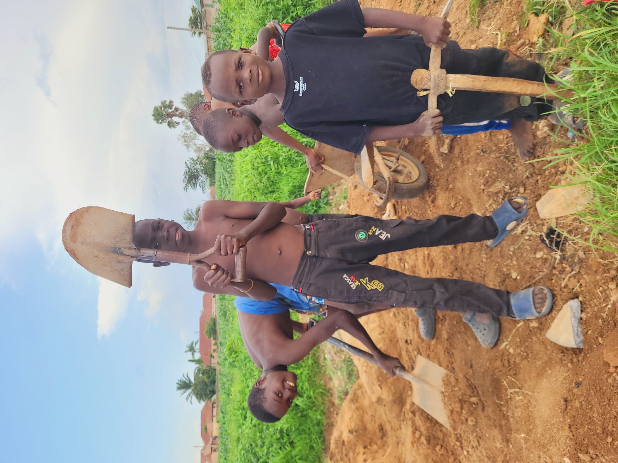 In Bukuru, Nigeria, four young boys are engaged in outdoor activity in a dusty, dirt-filled area with lush green vegetation in the background, suggesting a rural or semi-rural setting. The sky is a pale blue with scattered clouds. In the foreground, a boy wearing a black t-shirt and dark pants with a pickaxe that he is holding with both hands. To his left, another boy in dark pants is shoveling dirt. In the middle ground, a boy in blue shorts is looking at the camera while holding a shovel. Behind him, another boy in a red shirt is operating a wheelbarrow. The ground is covered in loose dirt and some green grass. In the background, there are buildings with red roofs and trees.