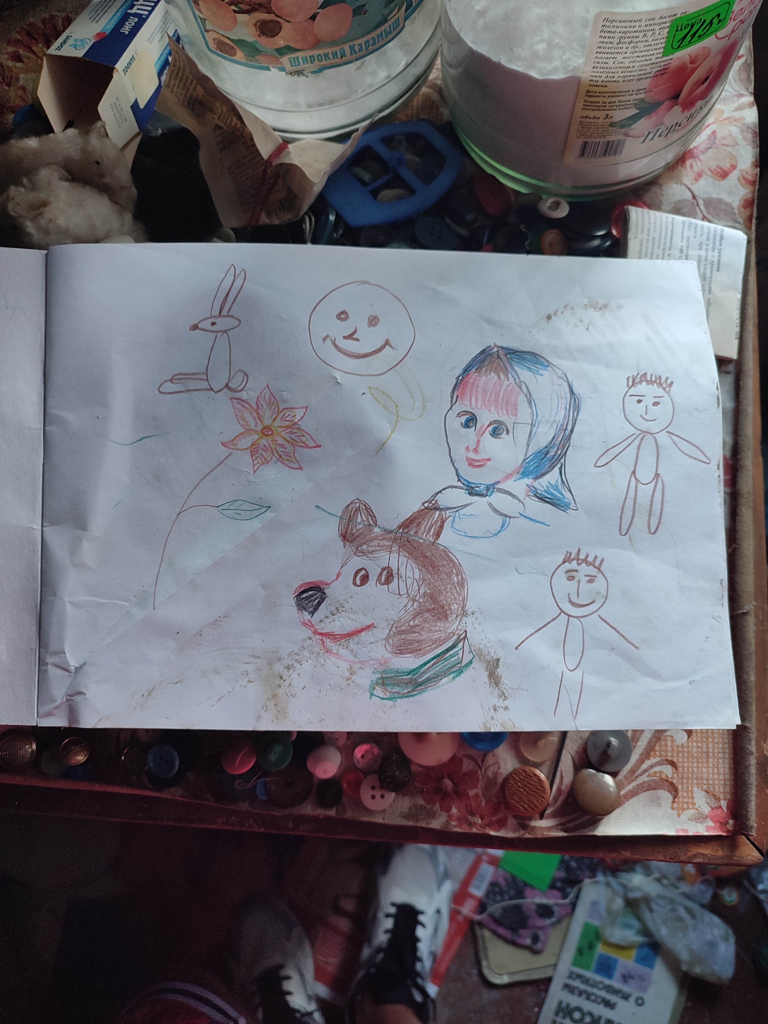 The image shows a child's drawing of a rabbit, a smiley face, a flower, a person with a headscarf, a dog, and two stick figures. The drawing is on white paper and appears to be done with colored pencils. The paper is creased and has some dirt smudges. The drawing is placed on a surface that looks like a table, with various buttons scattered around the edge. In the background, there are some containers and packaging, one of which appears to be a jar of jam. The scene is indoors, likely in a residential setting. The location is Gremyachinsk, Russia.