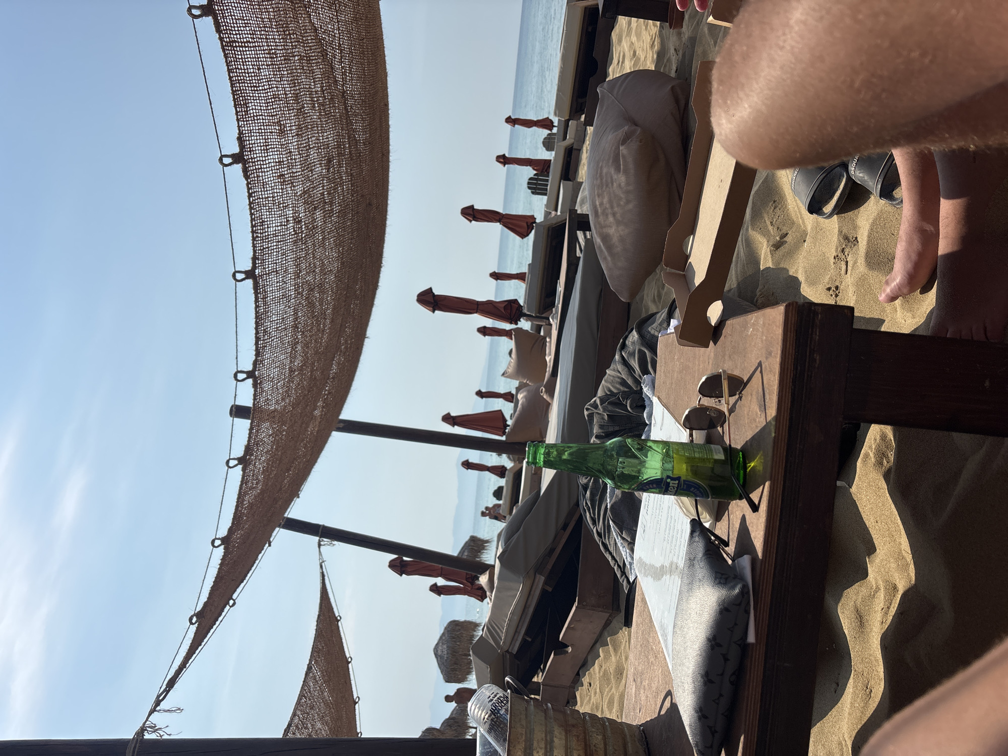 The image depicts a beach scene in Nea Peramos, Greece. In the foreground, a wooden table is set up on the sand, with a menu, sunglasses, a green glass bottle of Heineken, and a small dark pouch on it. To the left, a metallic bucket contains ice and what appears to be bottles of water. A person's legs and feet are visible in the bottom right and left of the frame, with a pair of dark slides near the feet on the sand. Behind the table, several beach loungers are arranged on the sand, some with cushions. Red beach umbrellas are open or closed in the background, along with thatched-roof sunshades further in the distance. The sea is visible beyond the loungers, with a clear blue sky overhead. The scene suggests a relaxed atmosphere at a beach bar or resort.