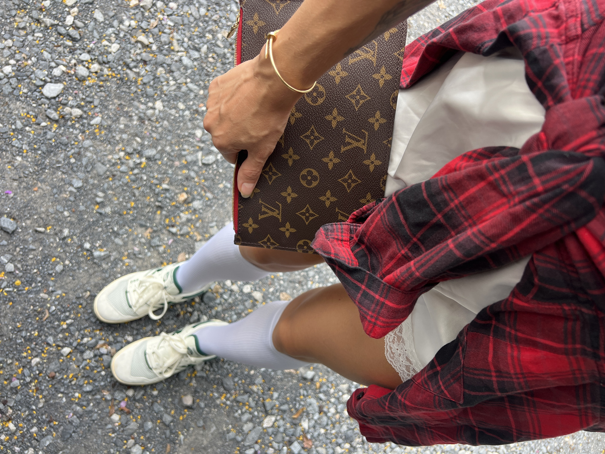 Here is a description of the image:
The image appears to be taken from an elevated perspective, focusing on a person's lower body and some accessories. The person is wearing a white slip dress with lace trim, a red and black plaid shirt, white knee-high socks, and white sneakers with green accents. They are holding a Louis Vuitton clutch in their left hand, adorned with a gold bangle.
The scene is set on what appears to be a paved surface, likely asphalt or concrete, scattered with small rocks or gravel. The location is identified only as Unknown, Unknown.