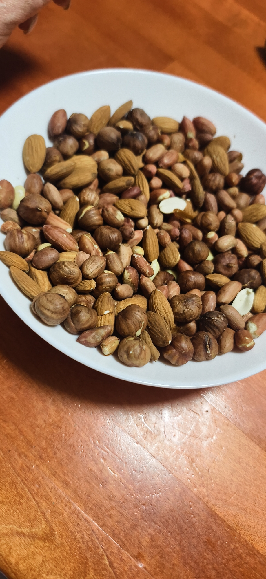 A white bowl is filled with a mix of nuts, including almonds, hazelnuts, and peanuts. The bowl is set on a wooden surface. The location is unknown.