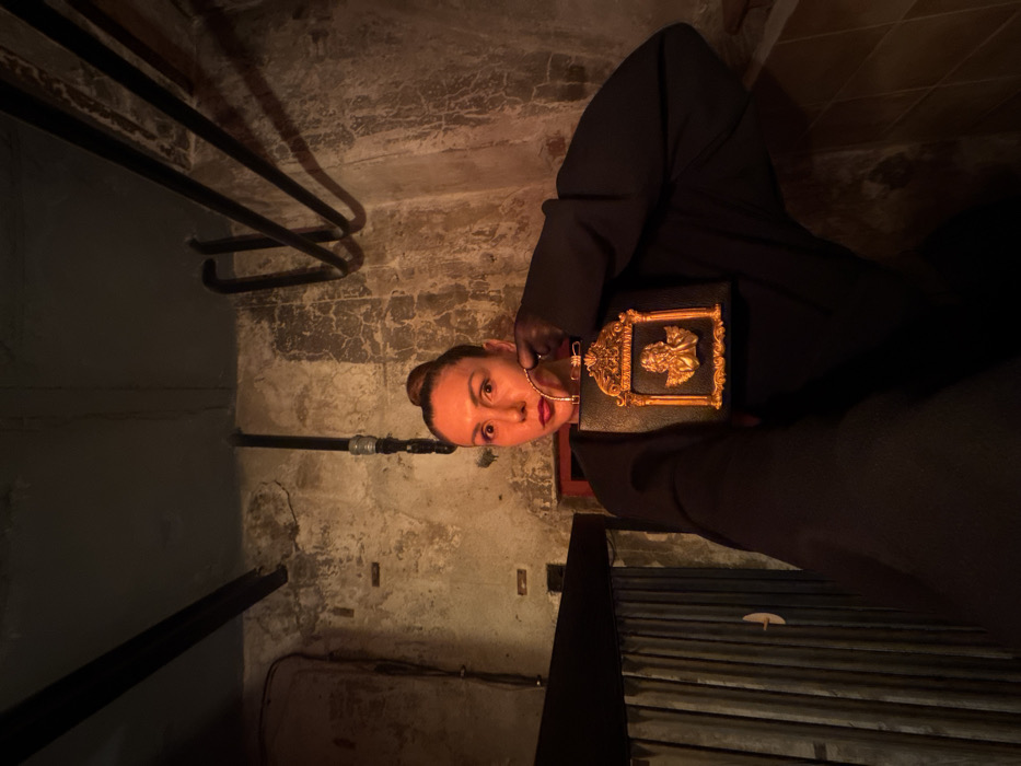 A female subject, estimated 30-50 years old, with dark hair in a high bun, red lipstick, and a wide-eyed expression, is depicted posing in a dimly lit, utilitarian interior space within Sofia, Bulgaria. She wears a dark, possibly black, long-sleeved garment with voluminous sleeves. Her right hand, clad in a dark, sheer glove, holds a black, rectangular handbag. The handbag features a prominent gold-colored ornate frame on its front, surrounding a raised gold-colored classical male bust sculpture, and is equipped with a metallic gold loop handle. The background consists of textured, aged light-colored walls with visible cracks and discoloration. Dark-colored utility pipes traverse the walls and ceiling. A dark horizontal beam and vertical corrugated paneling are visible on the left side of the frame. Warm, artificial lighting casts strong shadows, highlighting the textures of the environment.