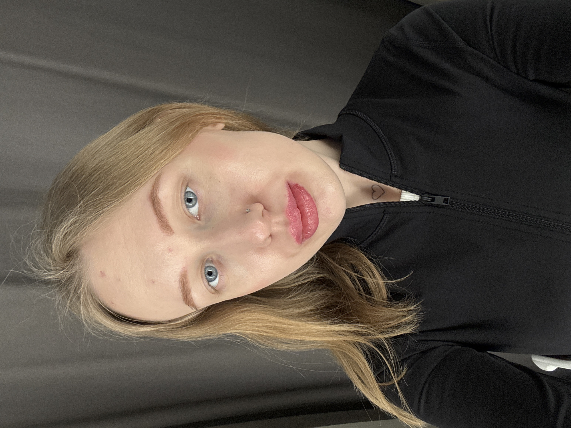 An adult female is depicted in a head-and-shoulder self-portrait, captured indoors in Rostov-on-Don, Russia. The subject possesses fair skin, blue eyes, and long, blonde hair. Her forehead exhibits several small, red cutaneous lesions. A silver-colored stud is visible in her nasal septum. Her lips are full and coated with a glossy pink-red substance. A small, black outline heart tattoo is present on her anterior neck, superior to the sternum. She is attired in a black, zippered athletic jacket, partially unzipped to reveal a white garment beneath. The background consists of a dark gray textile, exhibiting vertical folds, indicative of a curtain or drape. Lighting appears consistent with an internal light source, positioned anterior to the subject.
