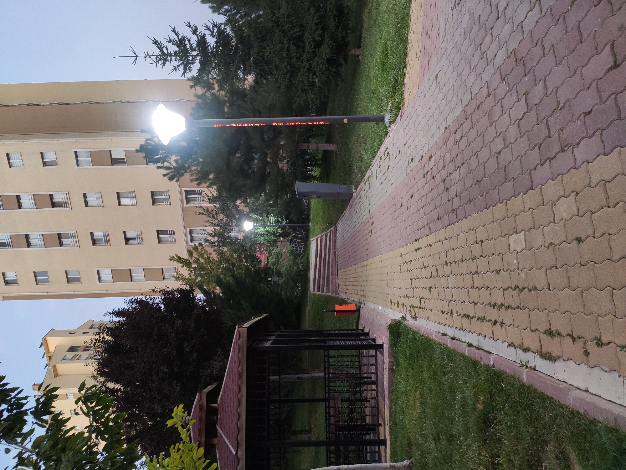 The image depicts a paved walkway in a park area with two tall, beige apartment buildings in the background. The walkway is constructed from interlocking bricks in shades of tan and reddish-brown. To the left of the walkway, there is a dark-colored gazebo with a maroon roof, featuring metal fencing and benches inside. Adjacent to the gazebo is a bright orange trash can. The ground to the left of the walkway is covered in green grass, and trees with dark green foliage are visible behind the gazebo. Further back, there are evergreens and the apartment buildings. A lamppost with a bright light is positioned on the right side of the walkway, and it has vertical red lettering on its pole that reads "ETİMESGUT BELEDİYESİ" (Etimesgut Municipality). The sky above is a pale, overcast blue, suggesting it is either early morning or late evening. The scene is tranquil and appears to be a public space within the city of Etimesgut, Turkey, specifically an urban park or residential complex. There are no people visible in the image.