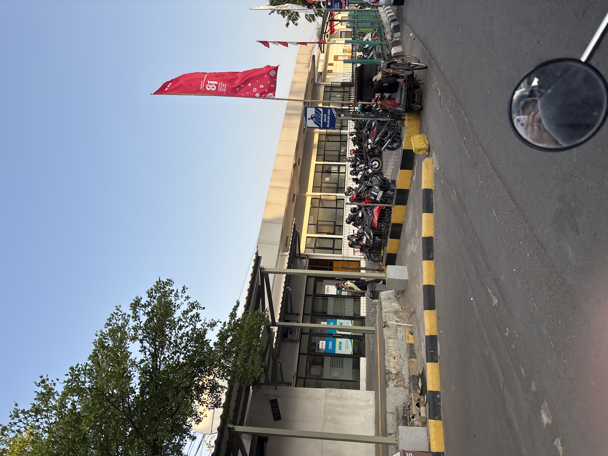 The image shows a street scene in Cirebon, Indonesia, likely near a transportation hub or public building. In the foreground, a motorcycle mirror reflects the hand holding a phone and part of the rider's arm and shoulder, indicating the photo was taken from a moving motorcycle. The road in front is asphalt, marked with yellow and black safety lines.
To the left, a building with a covered walkway and several ATMs is visible. A security guard in a light-colored uniform stands near the entrance. A large tree with green foliage partially obscures the building.
In the mid-ground, a parking area is filled with numerous motorcycles. A red flag with white polka dots and the number "81" is prominently displayed on a pole, likely representing a local event or celebration. Several smaller red and white flags are also visible. A three-wheeled vehicle, possibly a pedicab or becak, is parked nearby with a person seated in it.
The background consists of a low-rise building with large windows and a light-colored facade, suggesting it could be a government office, bank, or commercial establishment. The sky is clear and blue, indicating daytime, likely late afternoon due to the warm lighting. The overall impression is an active urban environment with a mix of infrastructure, vehicles, and people.